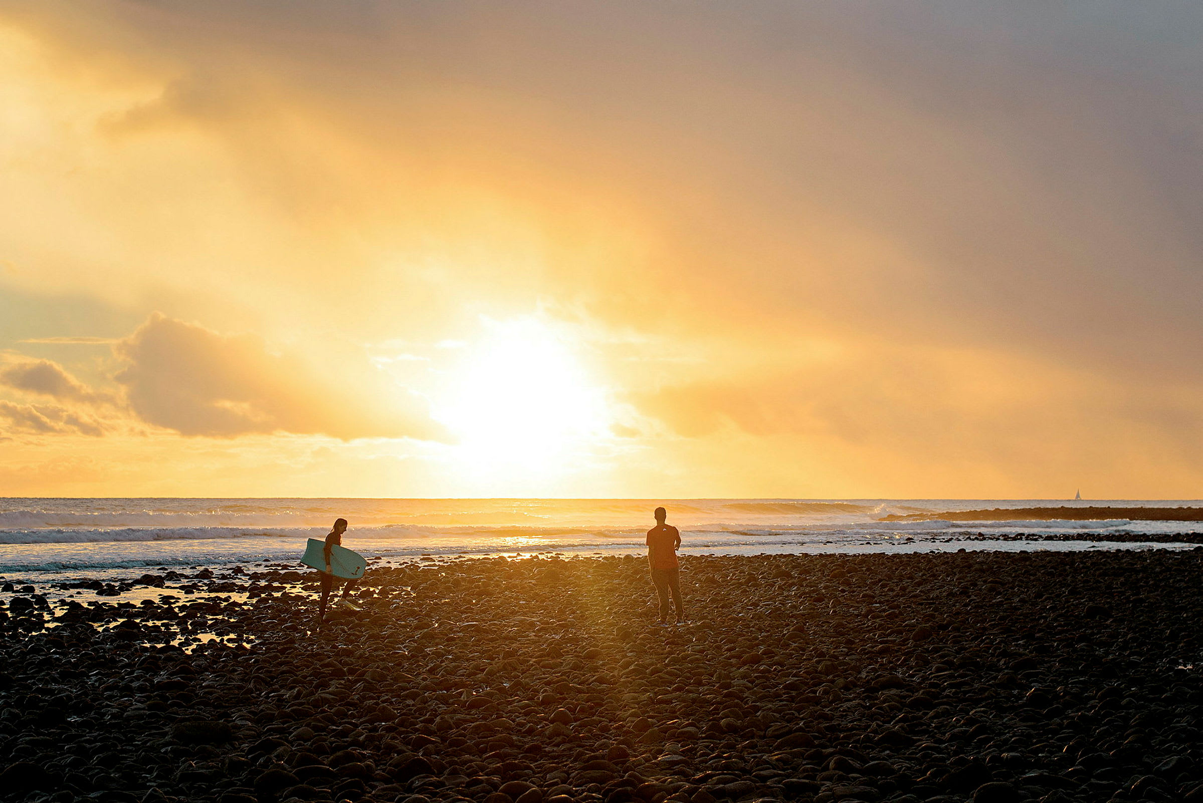 En solnedgång över havet med två personer på en stenig strand, den ena med en surfbräda. (AI-genererad text)