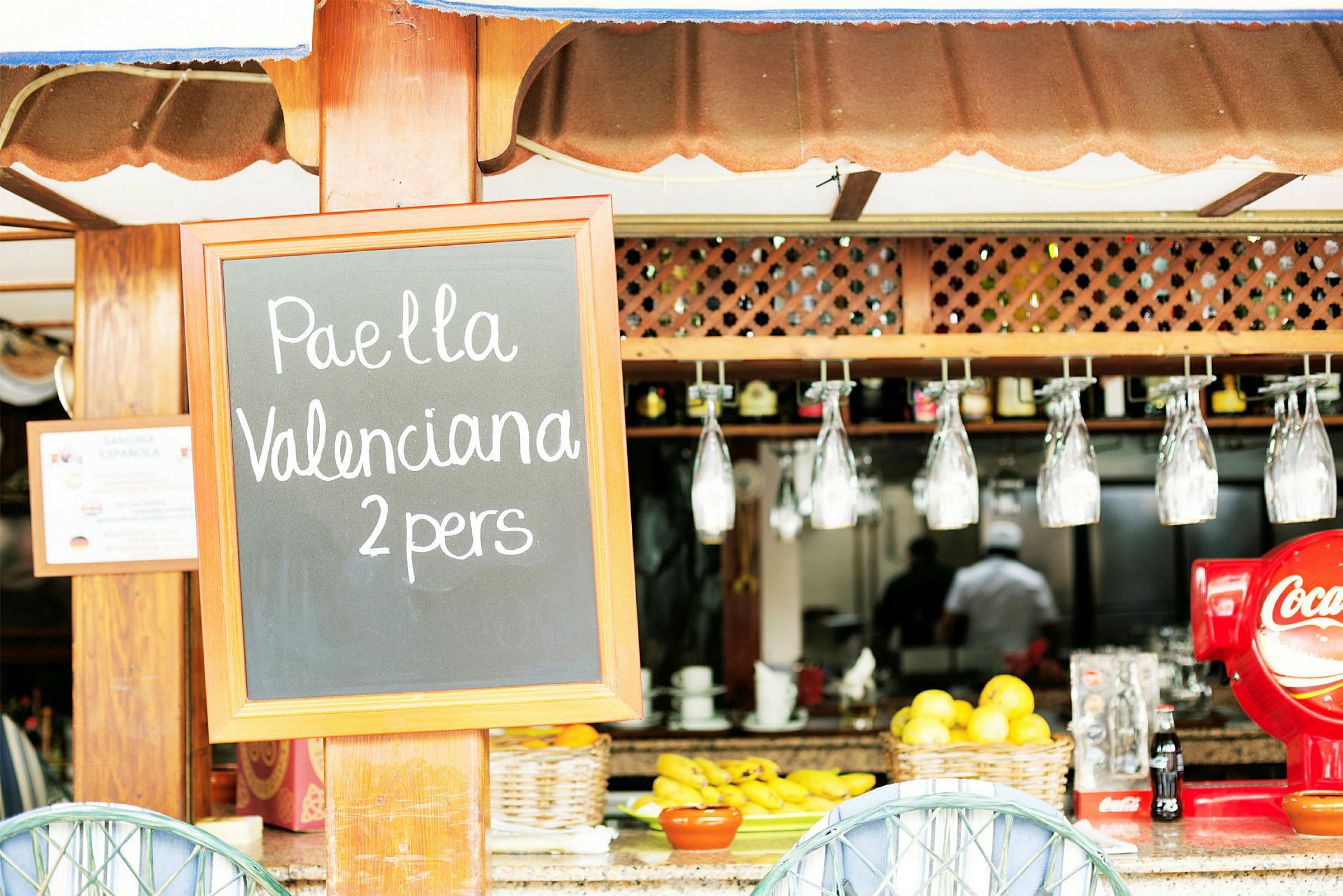 Skylt med texten 'Paella Valenciana 2 pers' framför en bar med frukt och glas. (AI-genererad text)
