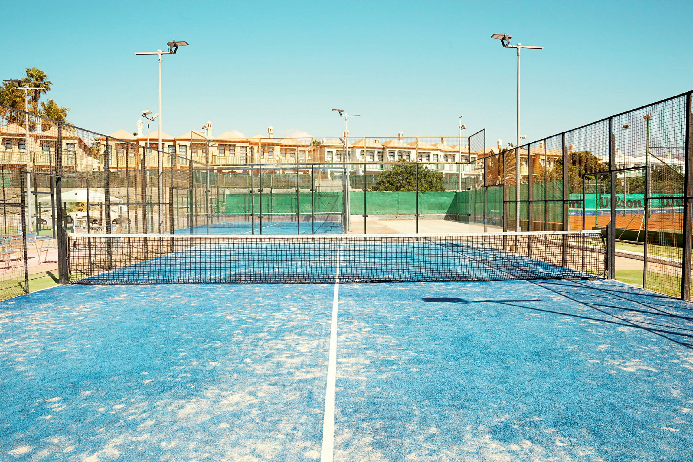 En padelbana med blått underlag och nät i mitten, omgiven av stängsel och byggnader i bakgrunden. (AI-genererad text)