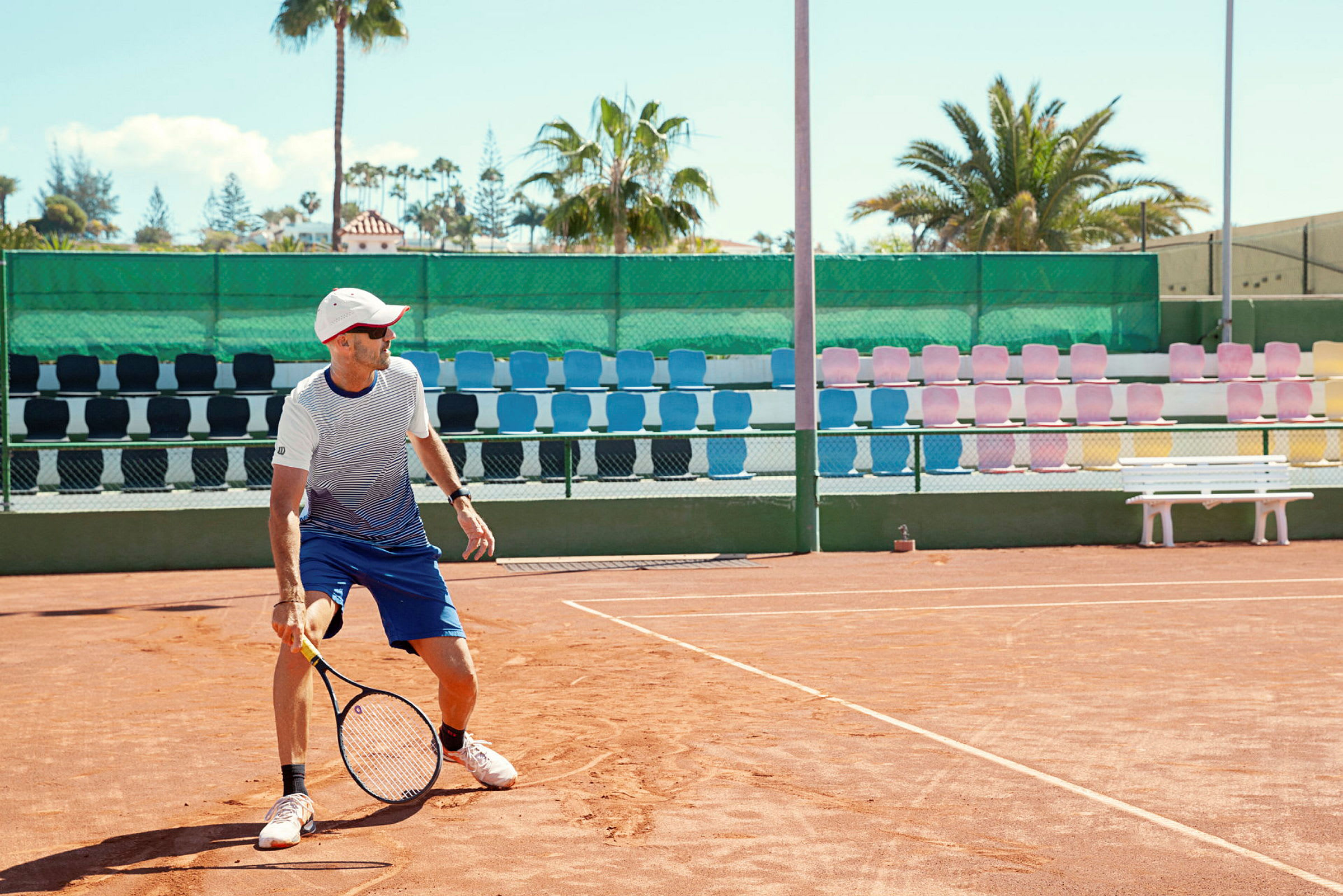En tennisplan med en spelare som håller en racket, omgiven av färgglada sittplatser och palmer i bakgrunden. (AI-genererad text)