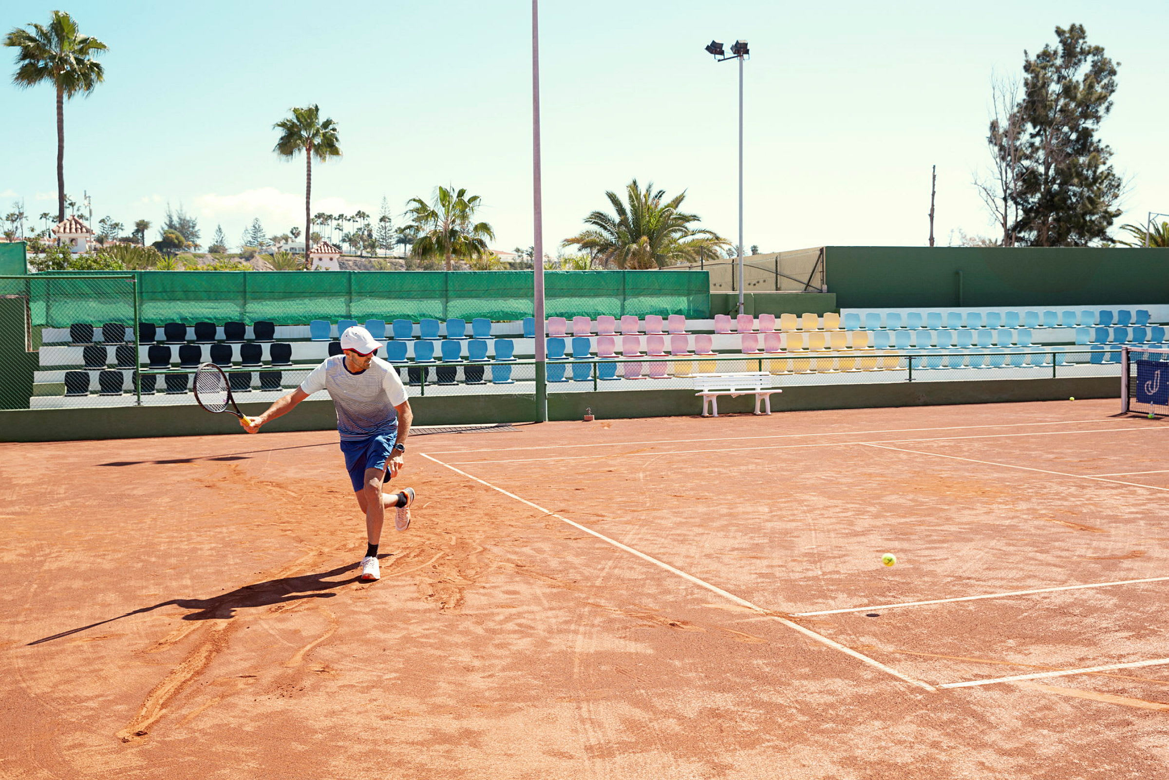 En person spelar tennis på en utomhusgrusplan med färgglada läktarstolar och palmer i bakgrunden. (AI-genererad text)