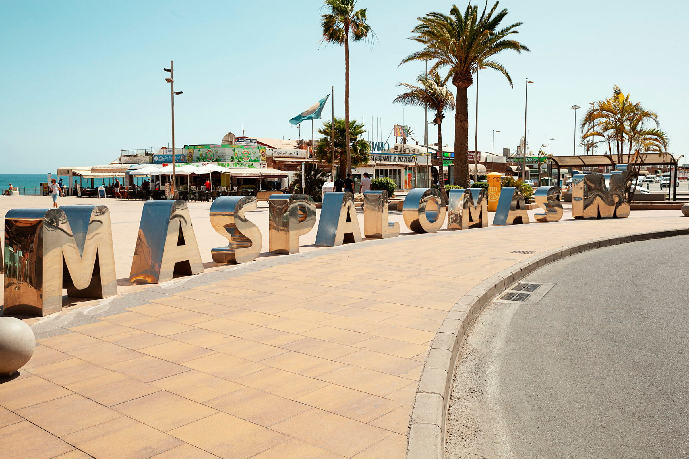Stora, blanka bokstäver som bildar ordet "MASPALOMAS" på en trottoar vid ett torg med palmer. (AI-genererad text)
