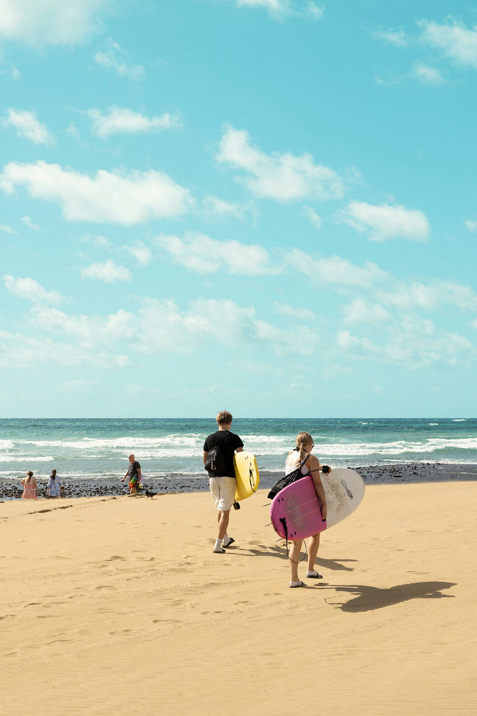 Två personer går mot havet på en sandstrand, bärande på surfbrädor. (AI-genererad text)