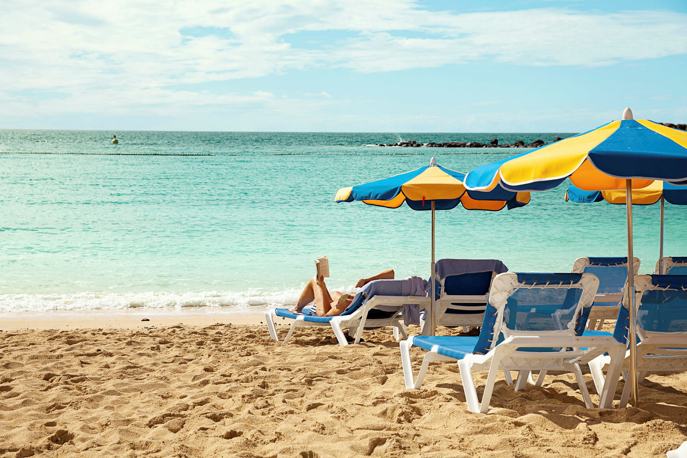 En strand med solstolar och parasoller, en person ligger och läser en bok. (AI-genererad text)