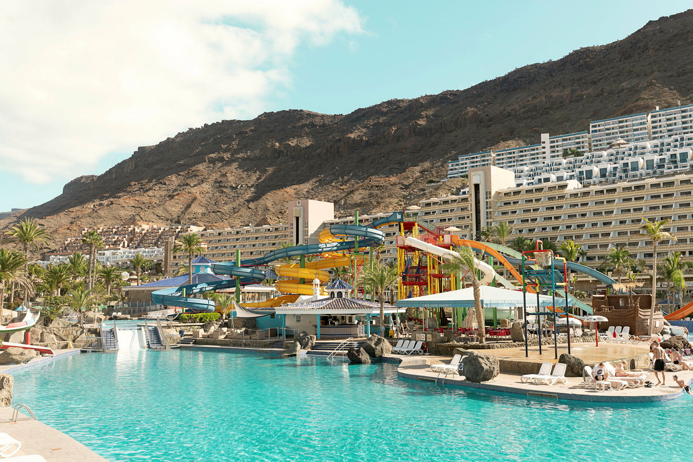 Stor pool vid ett hotell med färgglada vattenrutschbanor och berg i bakgrunden. (AI-genererad text)