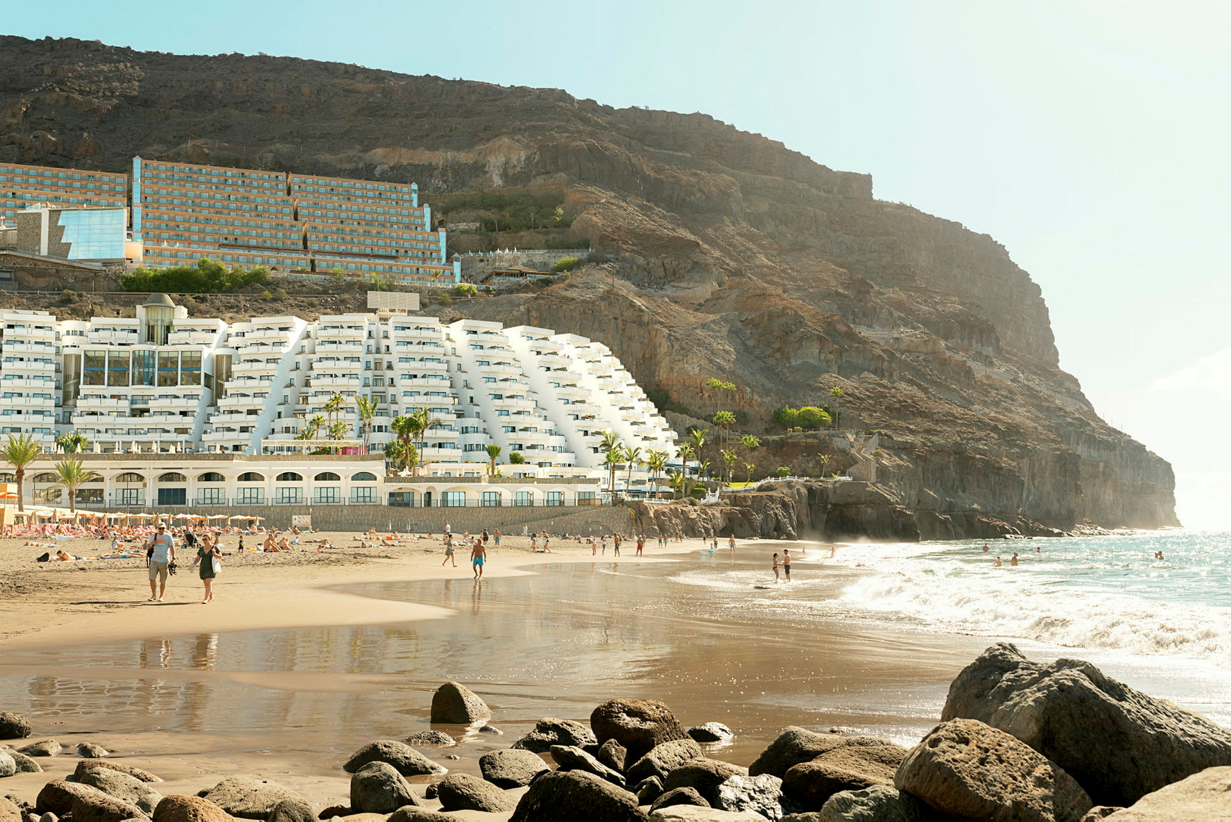 Strand med hotellbyggnad vid en sluttning och människor som rör sig längs sanden och vattnet. (AI-genererad text)