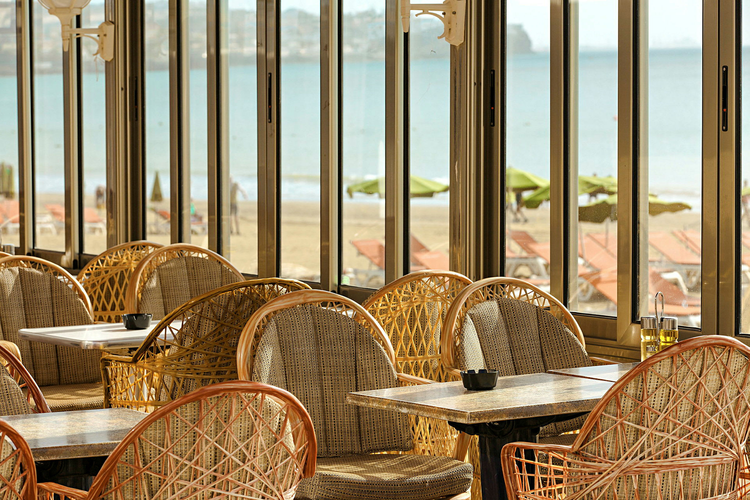 Utsikt från en restaurang med korgstolar och bord samt panoramafönster med utsikt över stranden och havet. (AI-genererad text)