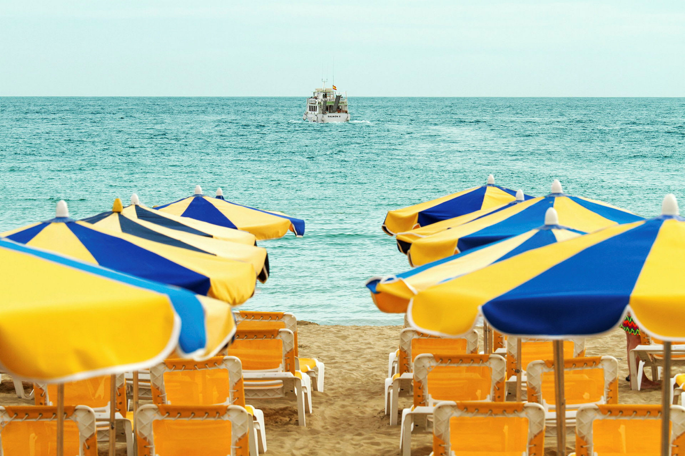 Solstolar och parasoller på en strand framför havet, med en båt i bakgrunden. (AI-genererad text)
