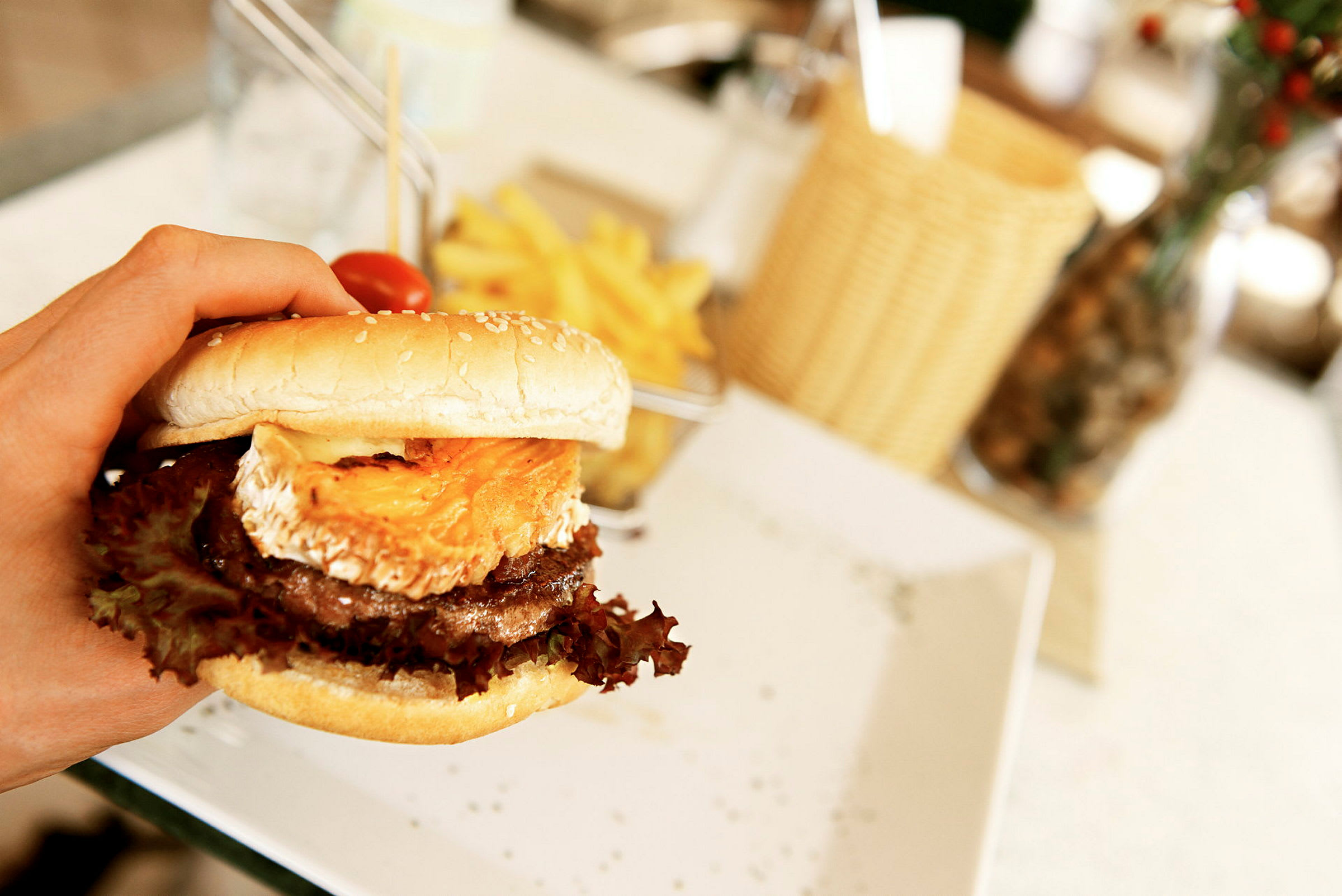 En hamburgare med ost och sallad hålls i en hand, med pommes frites på en tallrik i bakgrunden. (AI-genererad text)