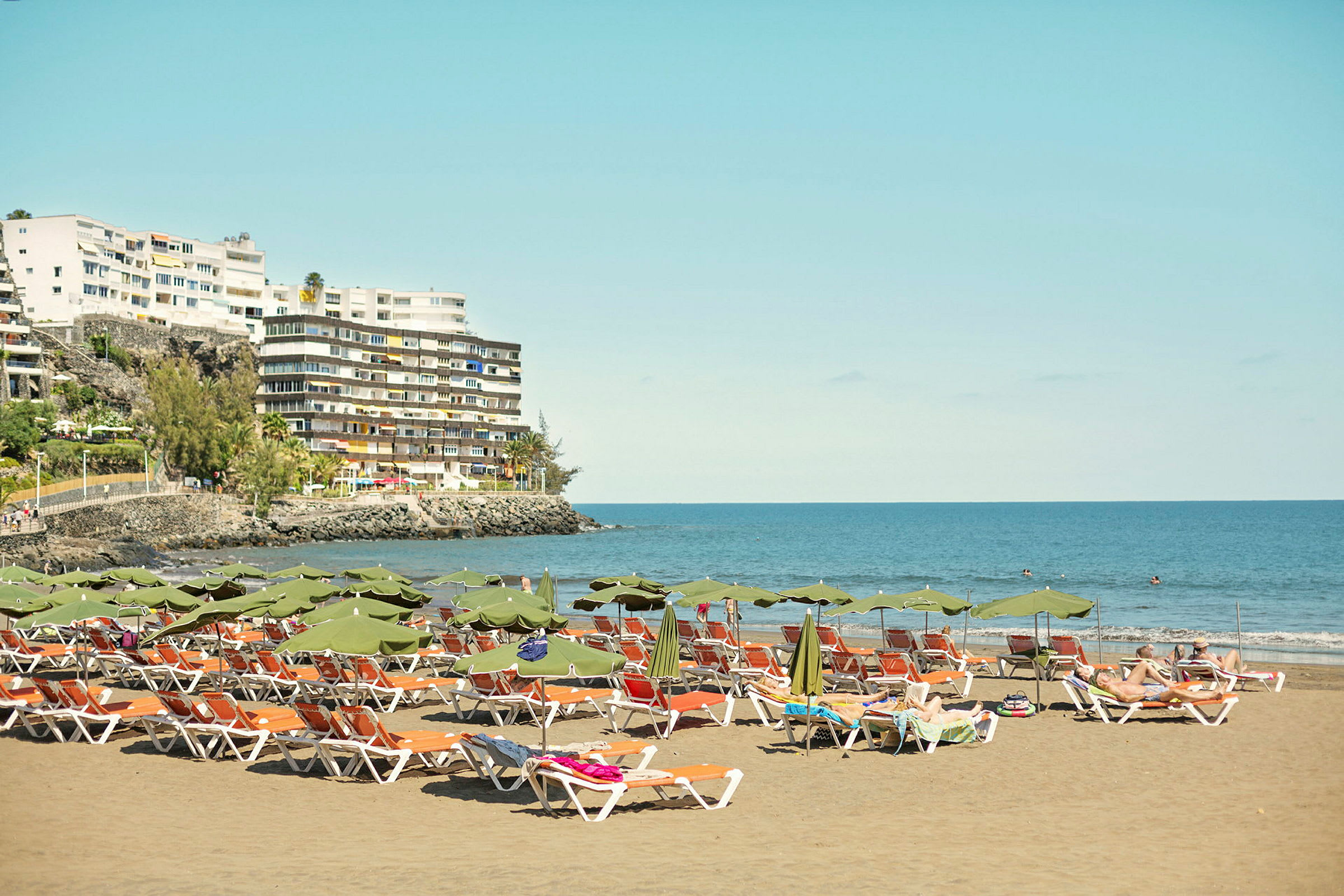 Strand med solstolar och parasoller, byggnader och hav i bakgrunden. (AI-genererad text)