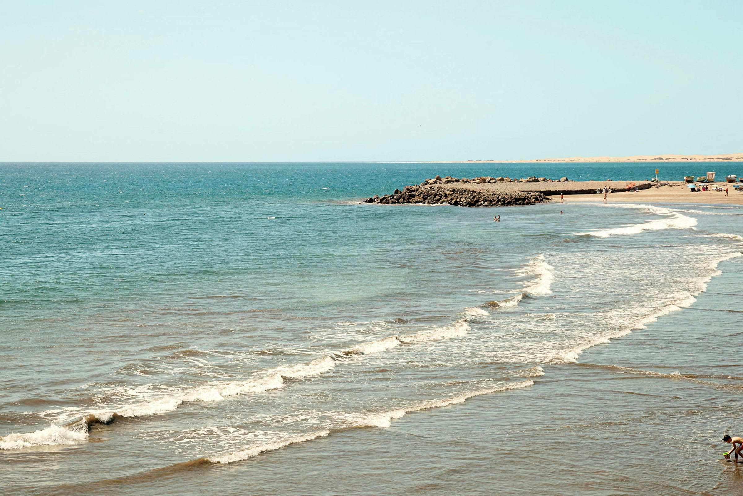 En strand med vågor som når land och människor som vistas vid vattnet. (AI-genererad text)