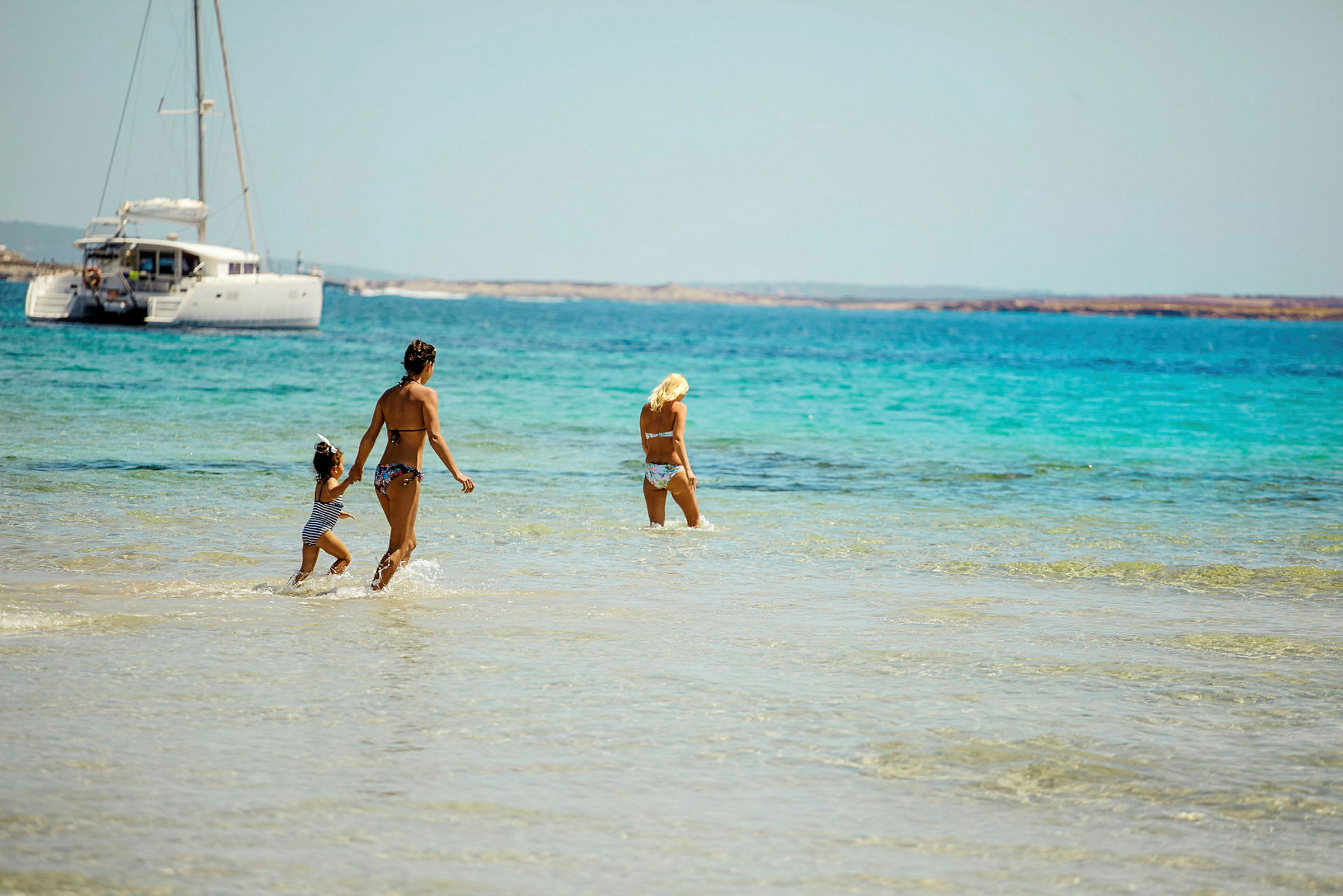 Tre personer går i grunt, klart vatten på en strand, med en segelbåt i bakgrunden. (AI-genererad text)