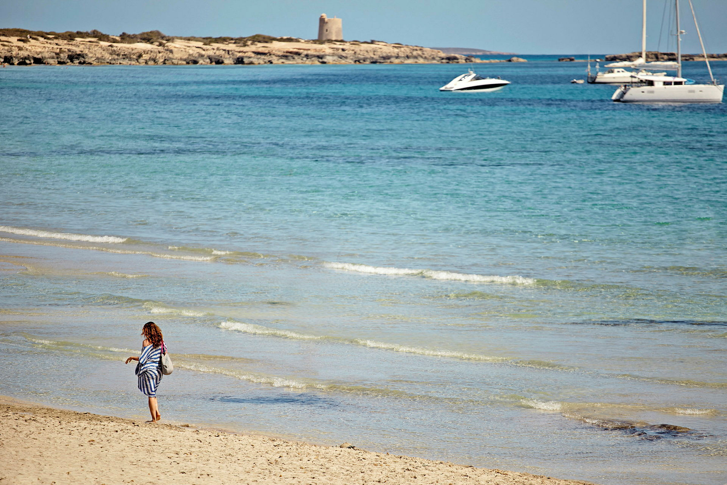 En person går längs en sandstrand vid havet med klara vågor. I bakgrunden syns en liten båt och en segelbåt ute på vattnet samt en klippa med en byggnad. (AI-genererad text)