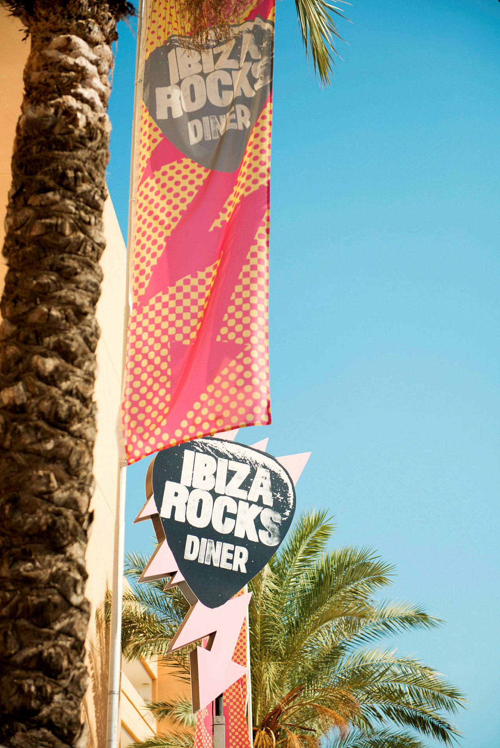 En skylt och banderoll för Ibiza Rocks Diner, omgivna av palmer och klar himmel. (AI-genererad text)