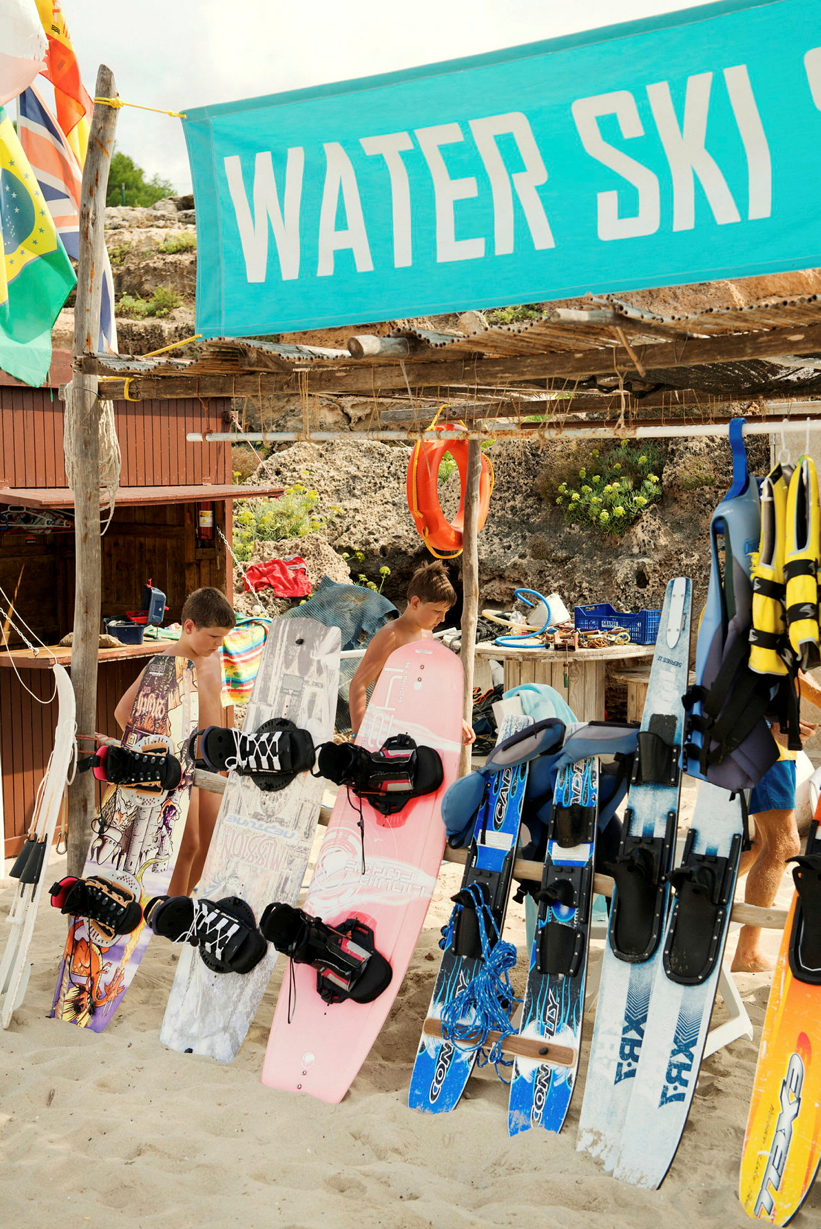 En samling av vattensportutrustning inklusive wakeboards och vattenskidor är uppställd på en strand nära en skylt som säger "Water Ski". (AI-genererad text)