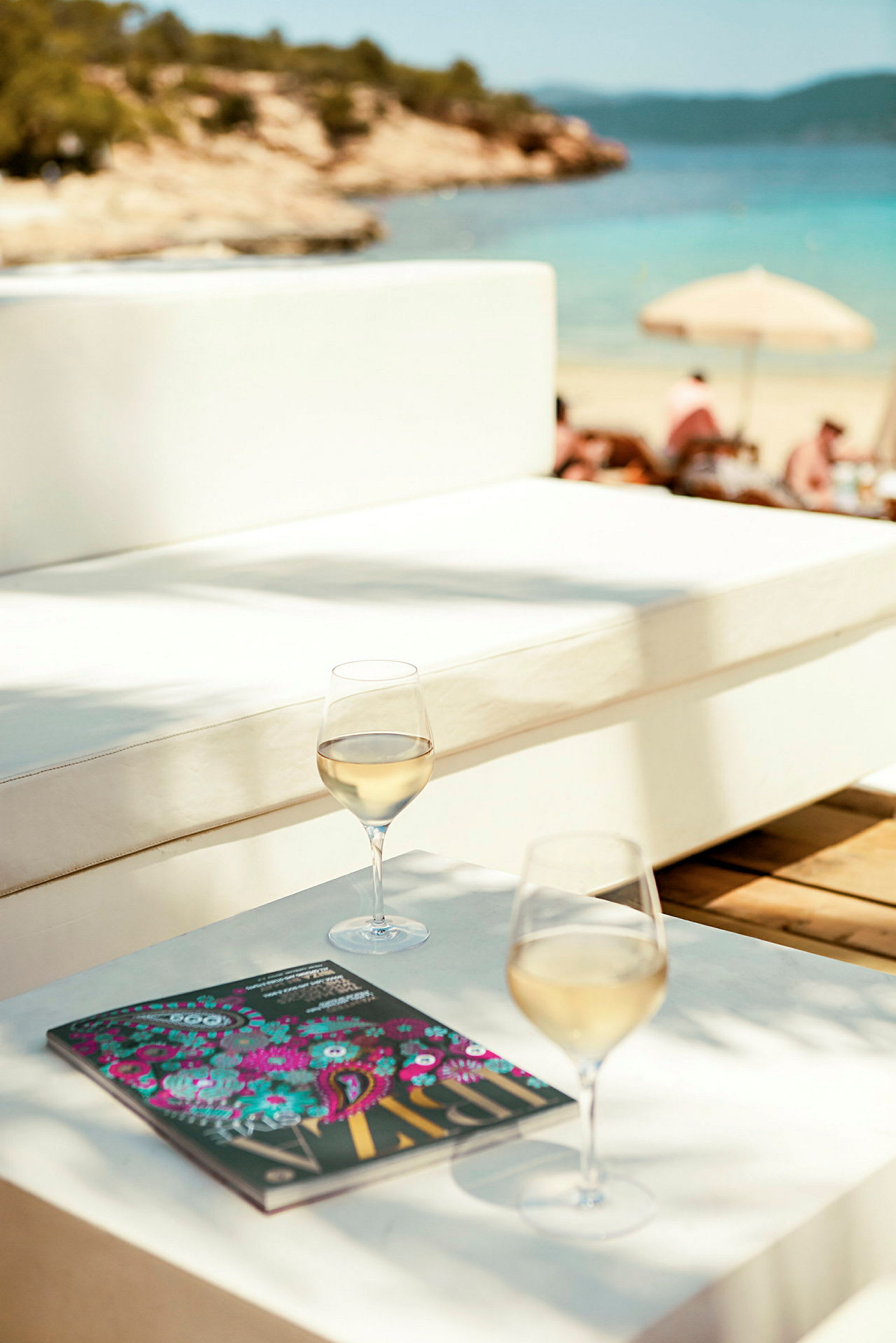 Ett bord med två glas vitt vin och en bok, med stranden och havet i bakgrunden. (AI-genererad text)