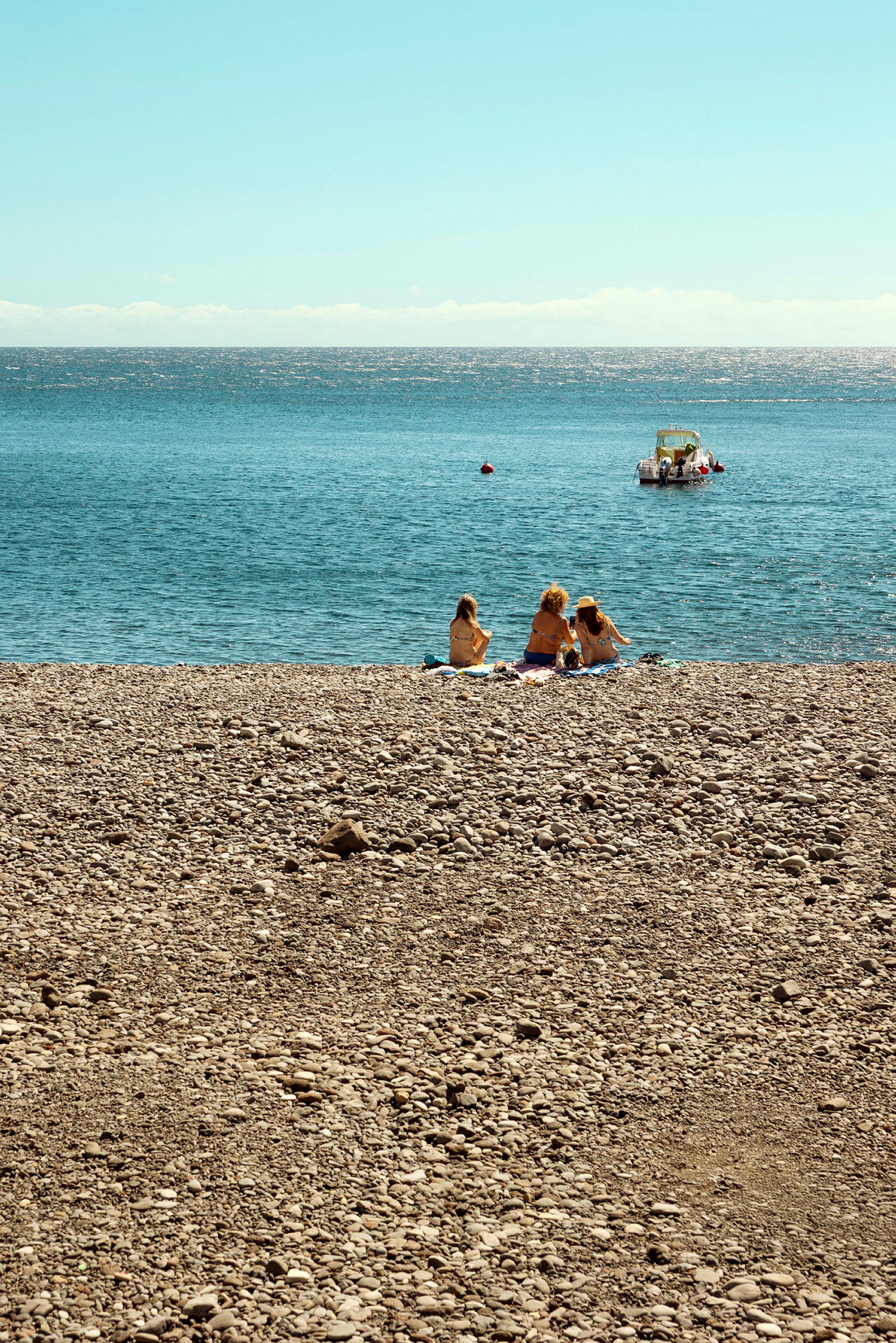 Tre personer sitter på en stenstrand med havet i bakgrunden och en båt längre ut på vattnet. (AI-genererad text)
