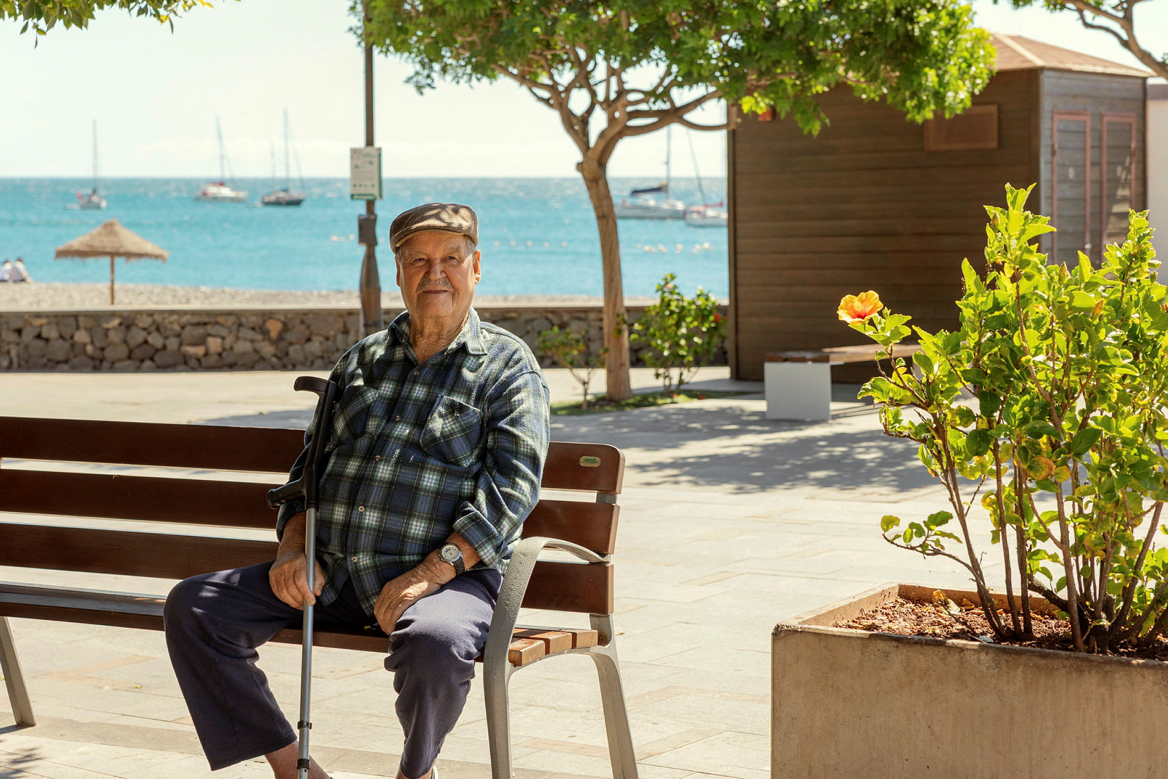 Person sitter på en träbänk vid en strandpromenad med havet och segelbåtar i bakgrunden. (AI-genererad text)