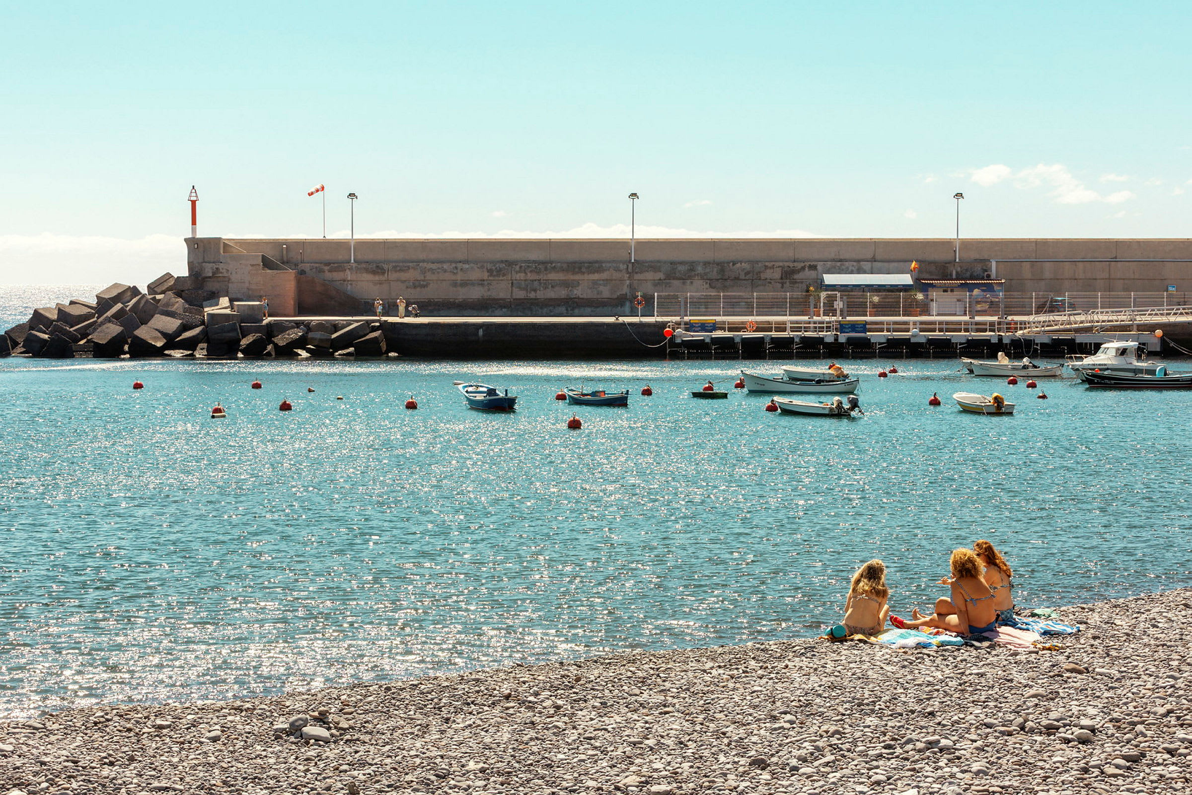 En strand med personer som sitter på handdukar vid vattnet, med små båtar förtöjda i hamnen. (AI-genererad text)
