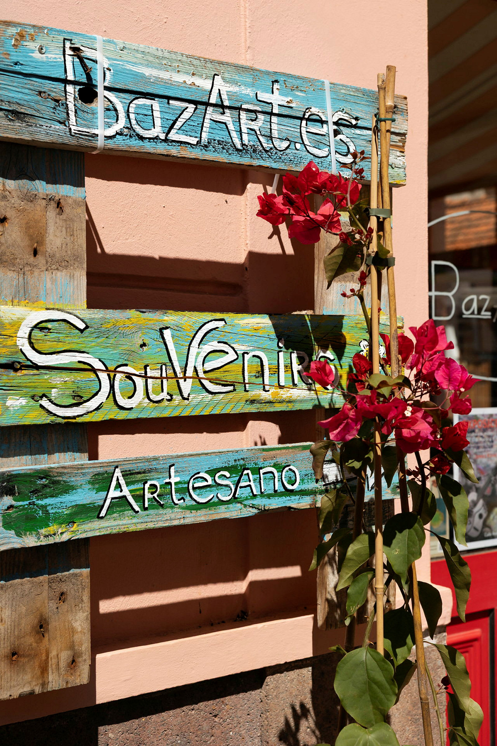 Träskylt med texten 'BazArt.es', 'Souvenirs' och 'Artesano' omgiven av röda blommor. (AI-genererad text)