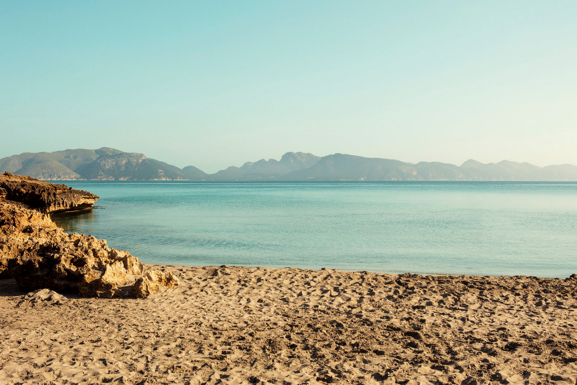 En sandstrand med klippor vid havet och berg i bakgrunden. (AI-genererad text)