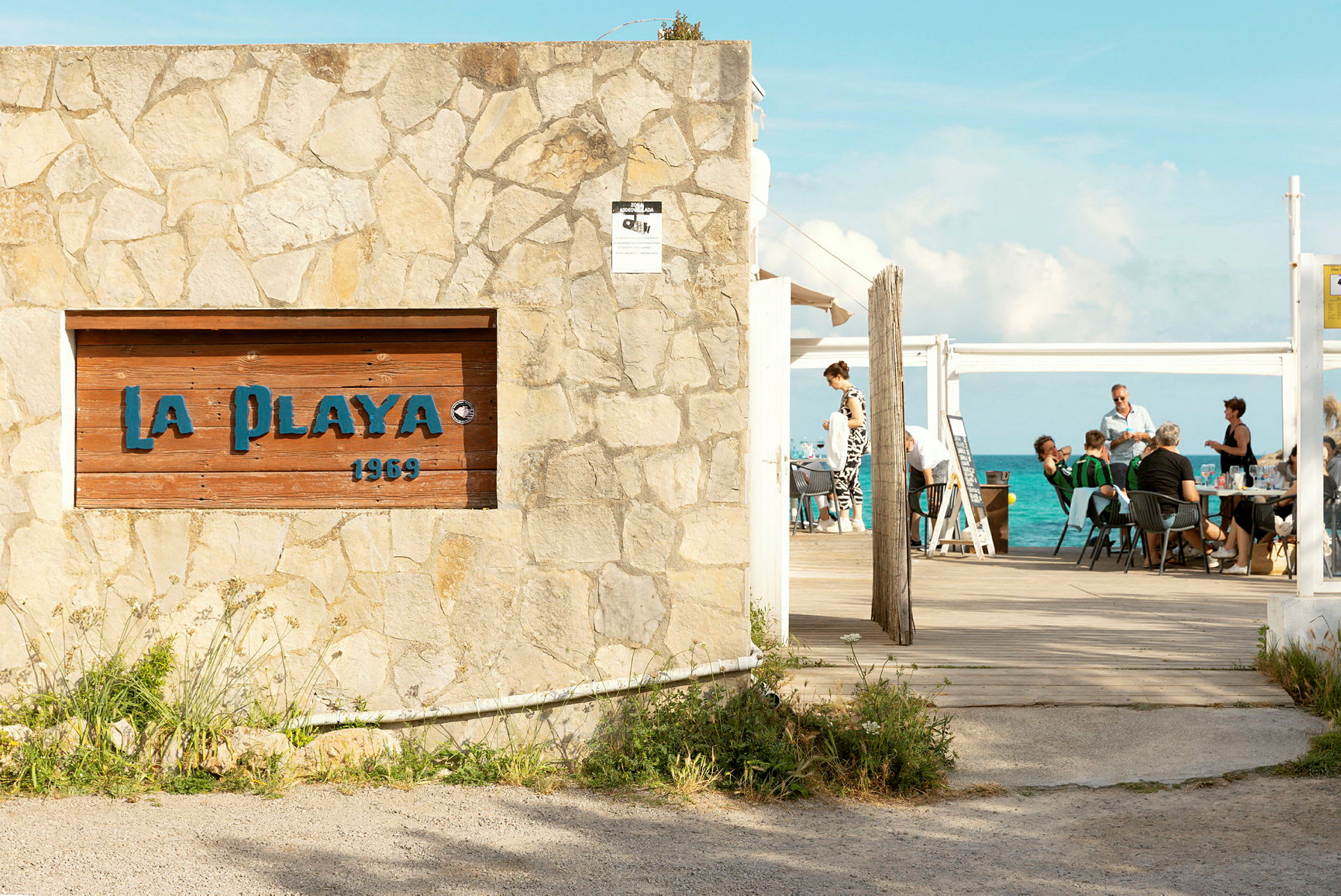 En stenmur med en skylt som har texten 'La Playa 1969' framför en utomhusservering med havet i bakgrunden. (AI-genererad text)