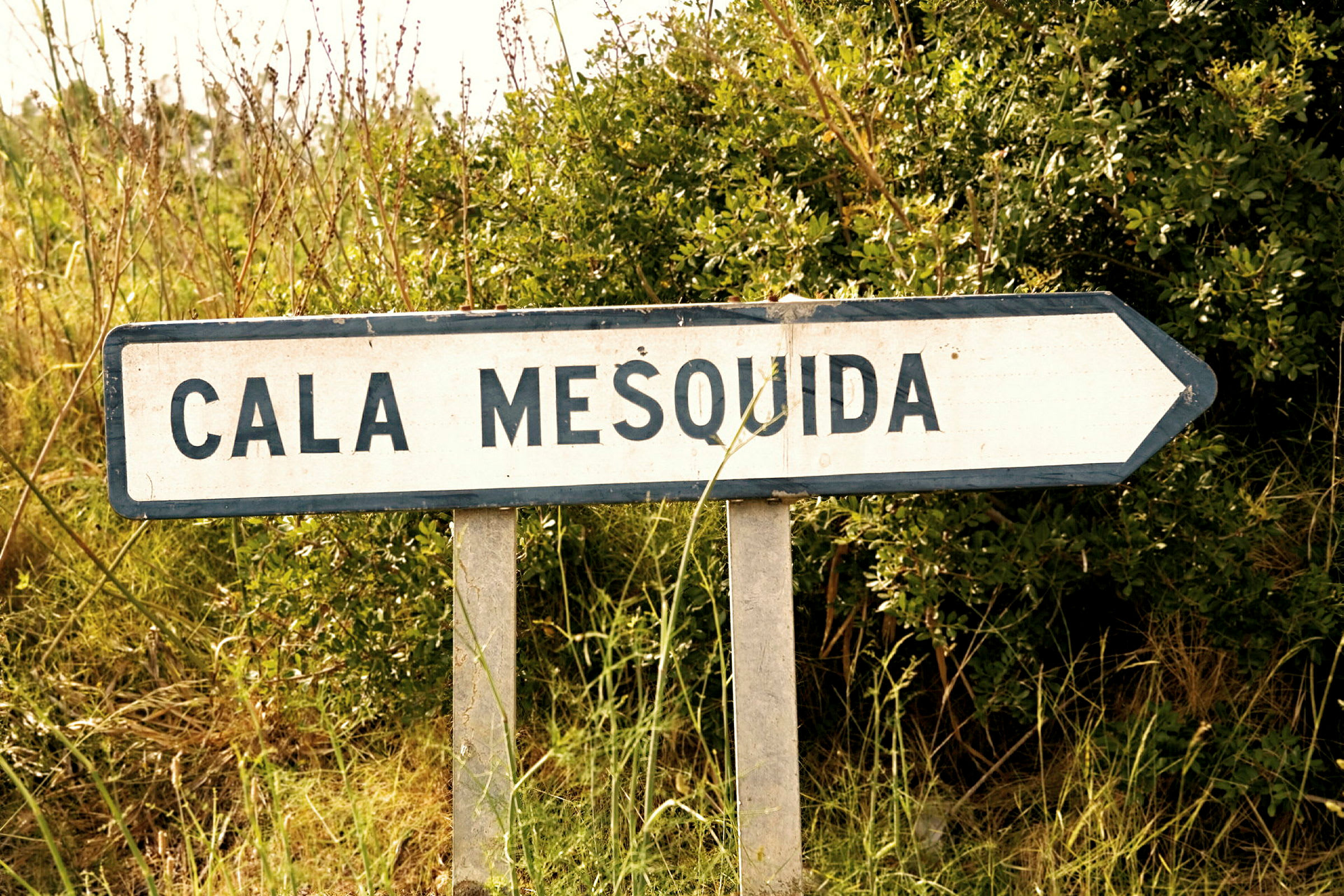 En vägskylt med texten "Cala Mesquida" pekar åt höger, omgiven av grönska. (AI-genererad text)