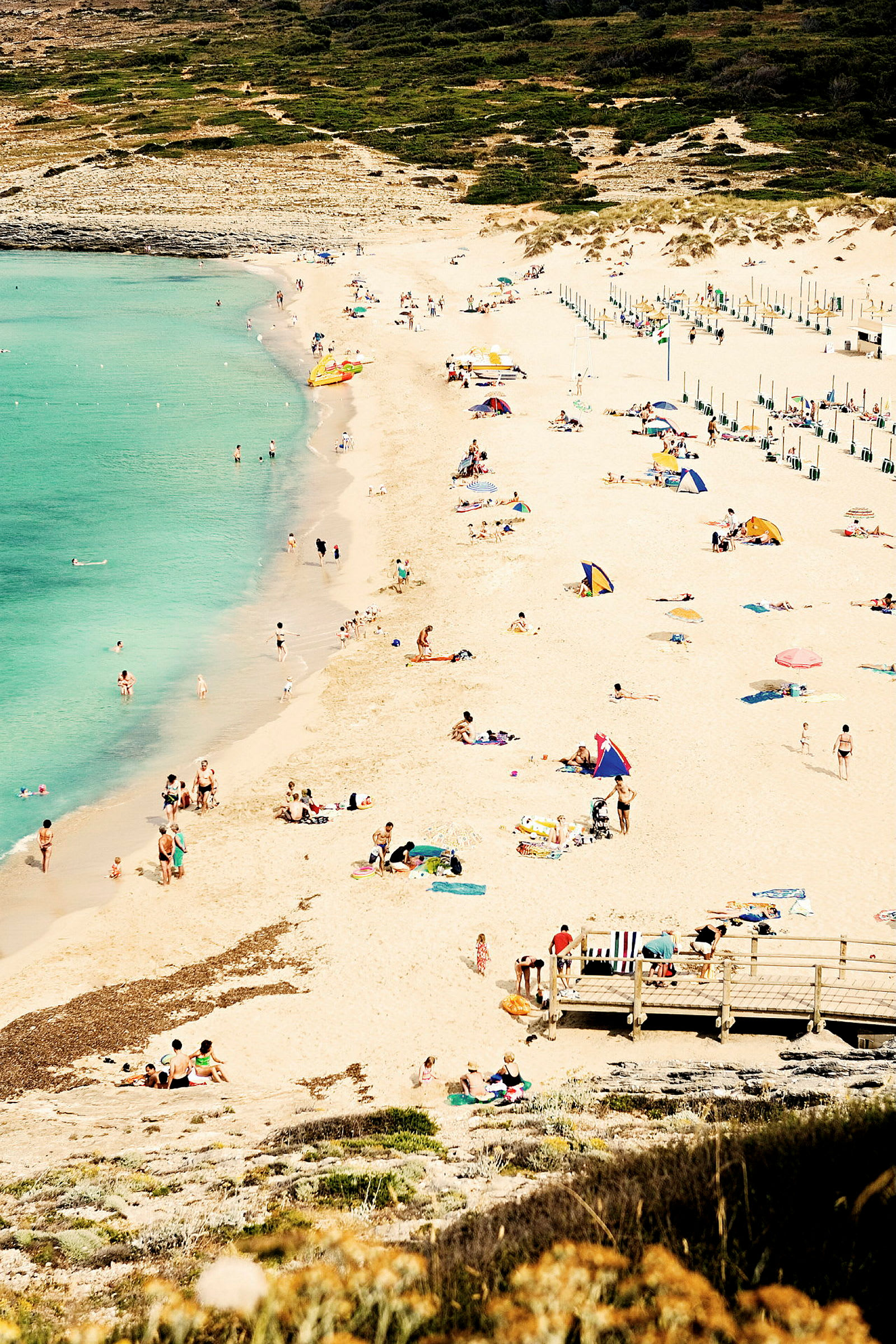 En sandstrand med solbadande människor och turkost vatten i bakgrunden. (AI-genererad text)