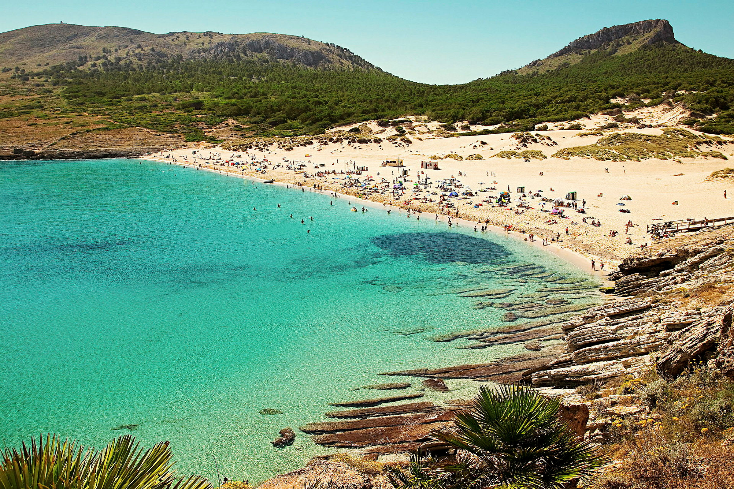 En sandstrand med kristallklart vatten och människor som solar och badar. Skogklädda berg syns i bakgrunden. (AI-genererad text)