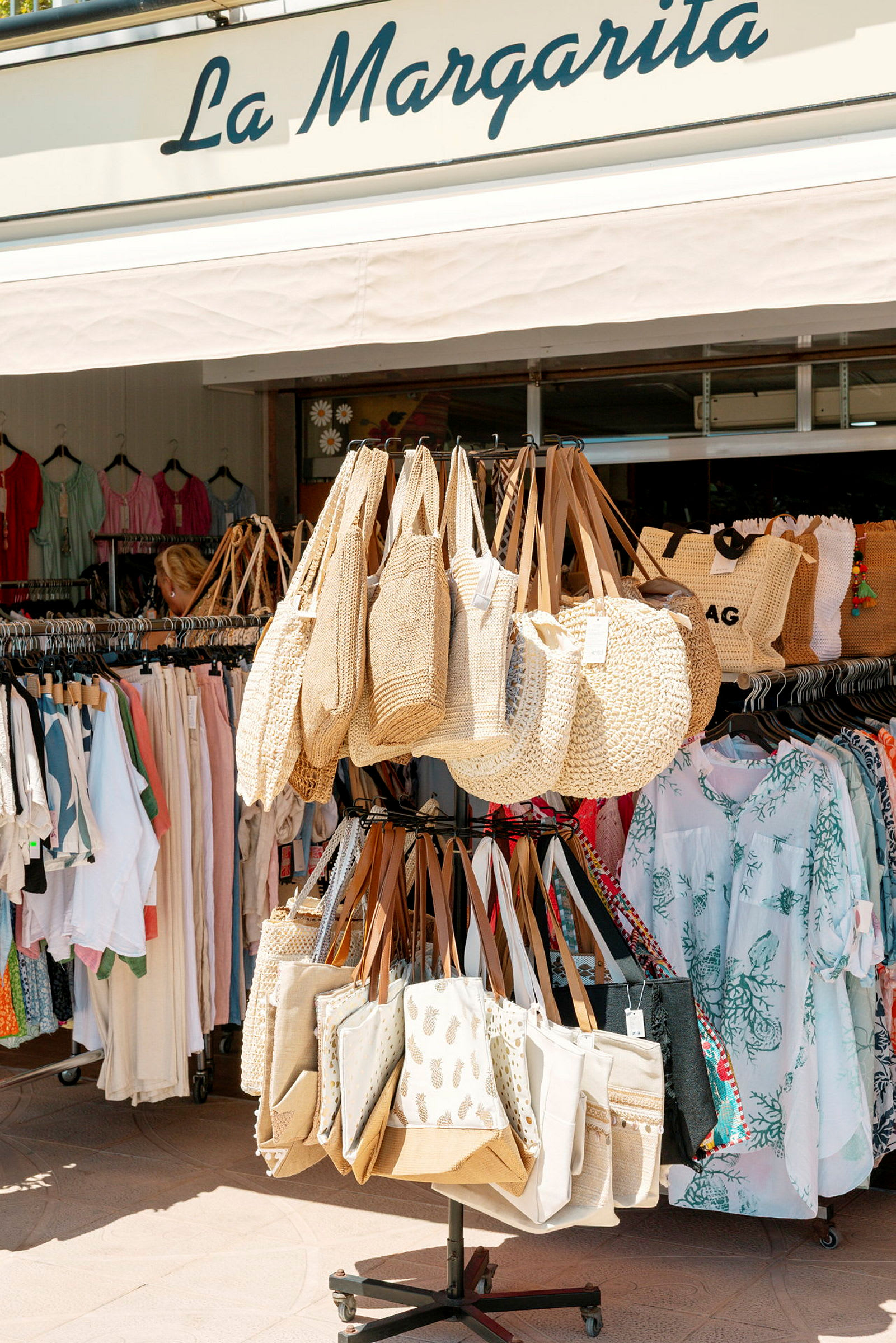 Ett stativ med strandväskor framför en butik som säljer kläder och accessoarer. (AI-genererad text)