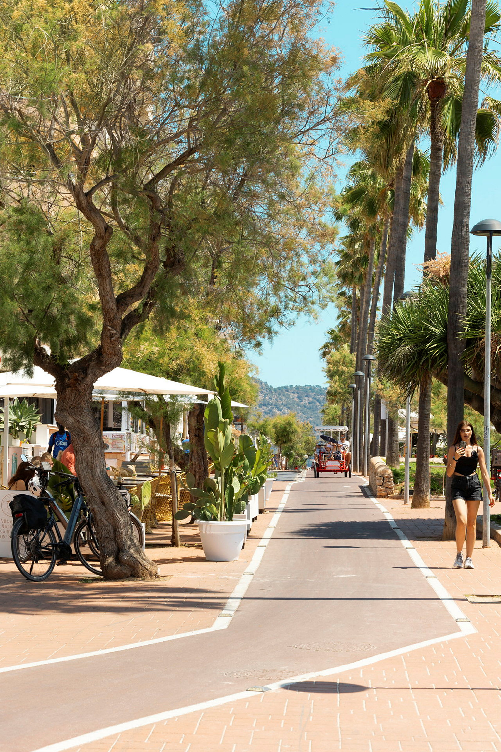 En promenadkantad gata med träd och palmer, cyklar och serveringar på vänster sida samt personer och ett turisttåg i fjärran. (AI-genererad text)