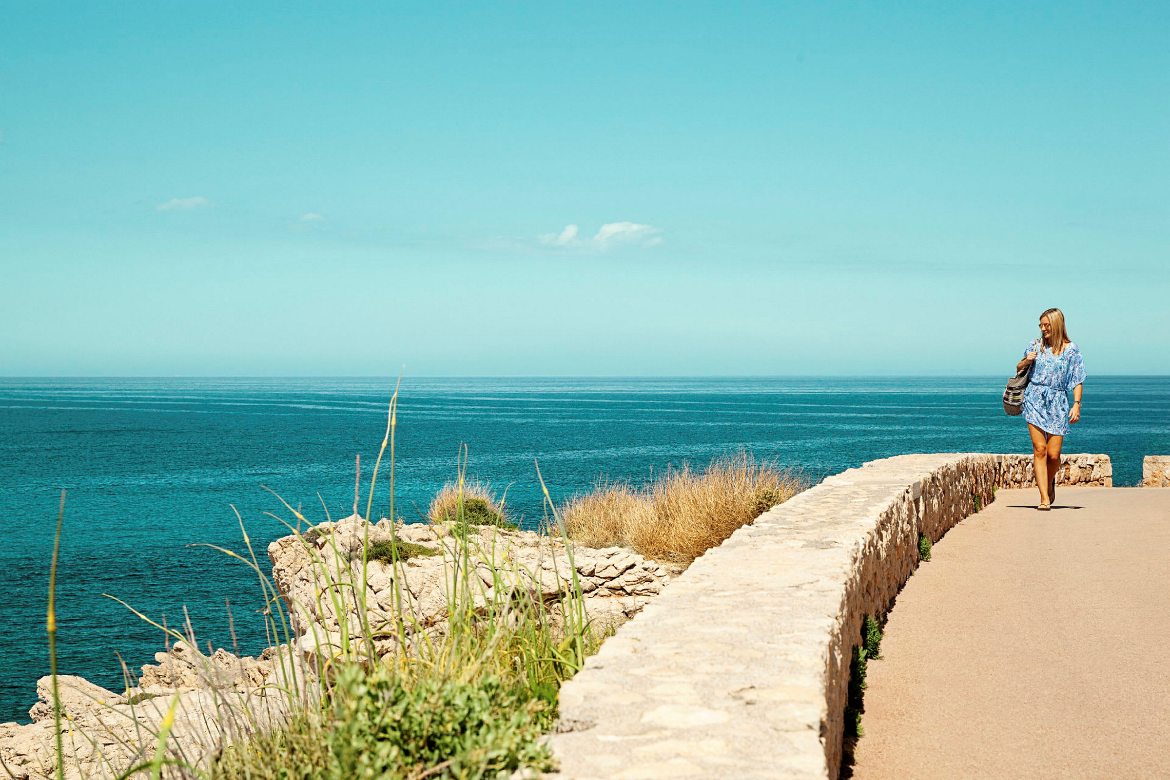 Kvinnlig person promenerar vid en stig som vetter mot havet, med sten och vegetation i förgrunden. (AI-genererad text)