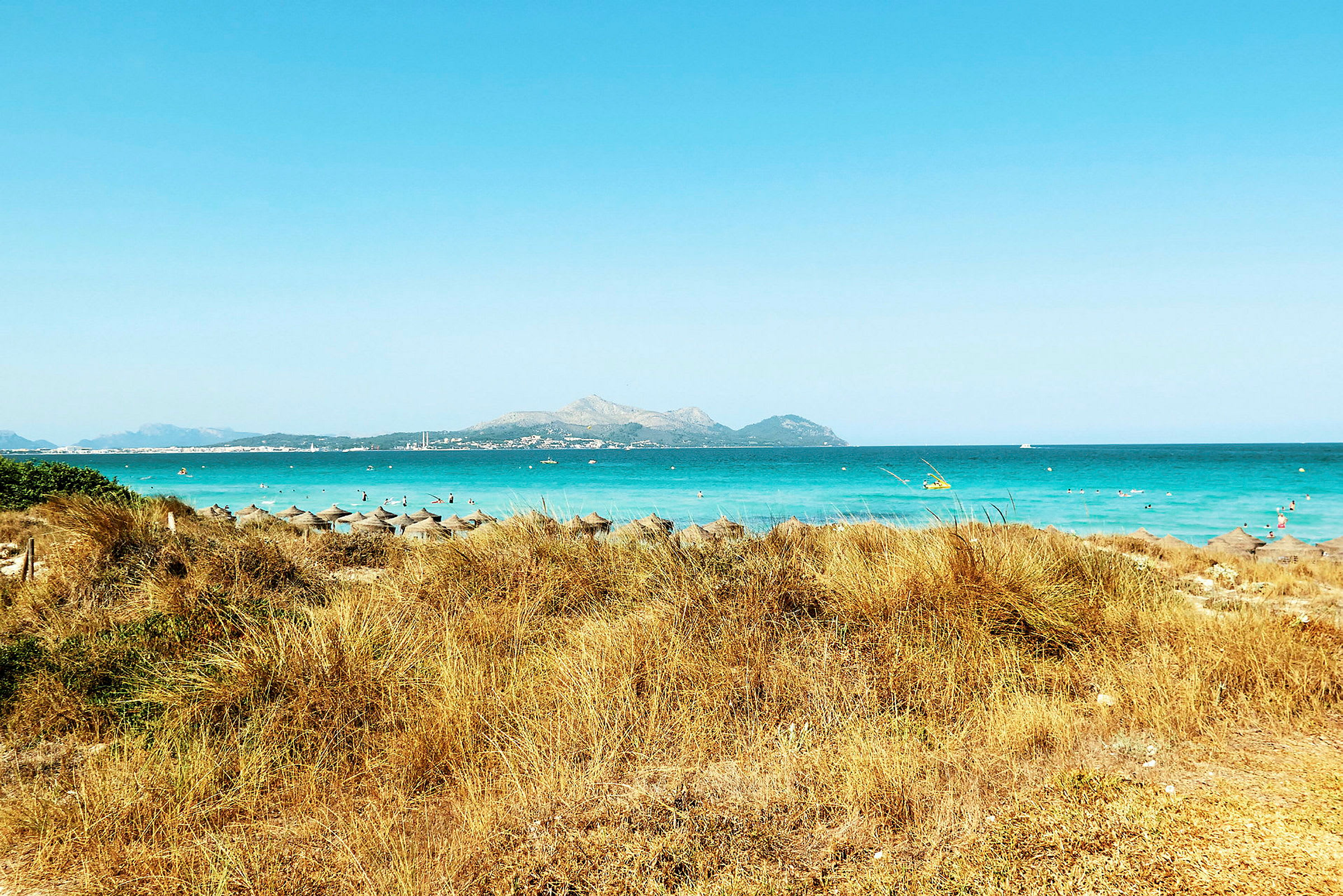 En strand med solparasoller, turkost vatten och berg i bakgrunden. (AI-genererad text)