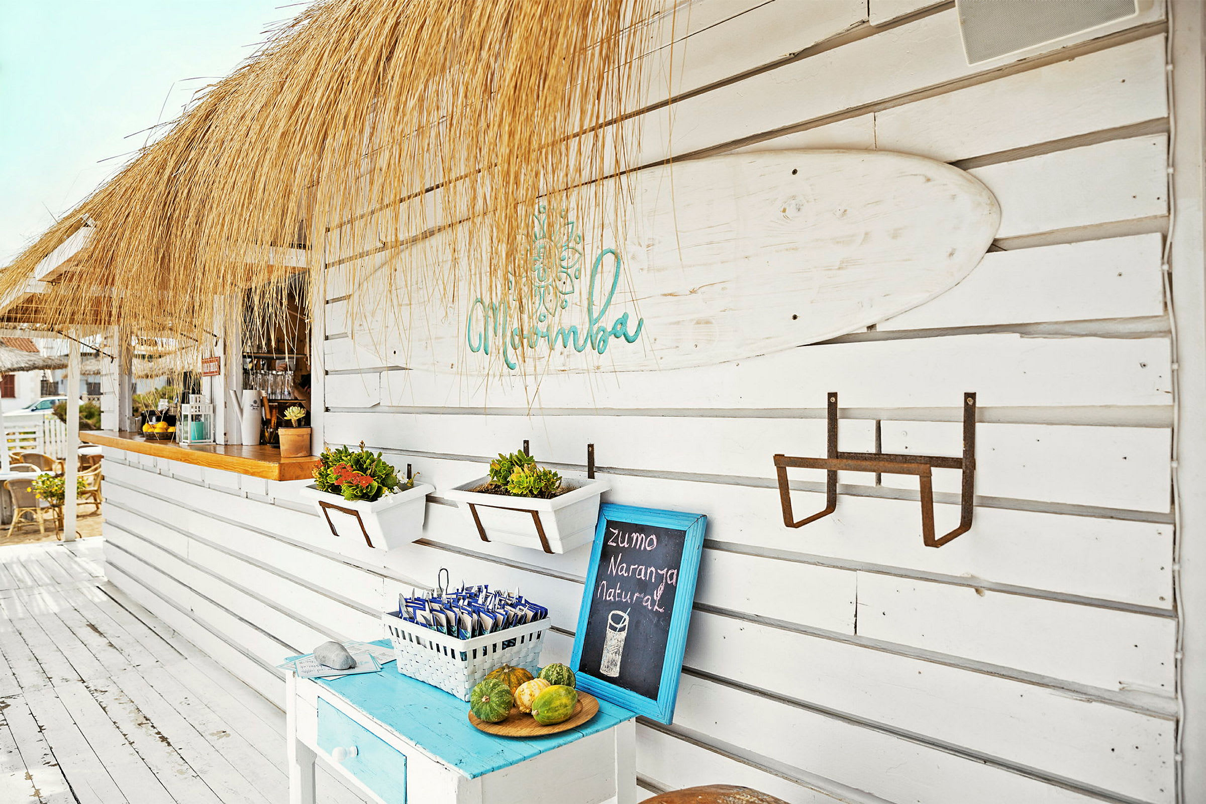 En strandbar med halmtak och vita träväggar dekorerad med surfbräda, blomlådor och en skylt. (AI-genererad text)