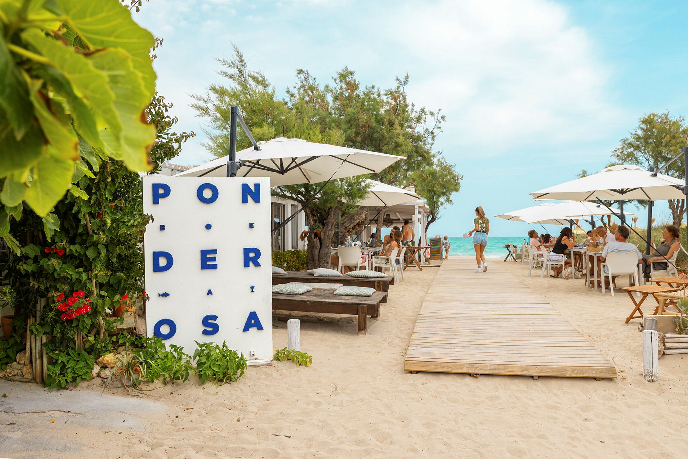 En restaurang vid stranden med bord och parasoller, en trägång leder mot vattnet. (AI-genererad text)