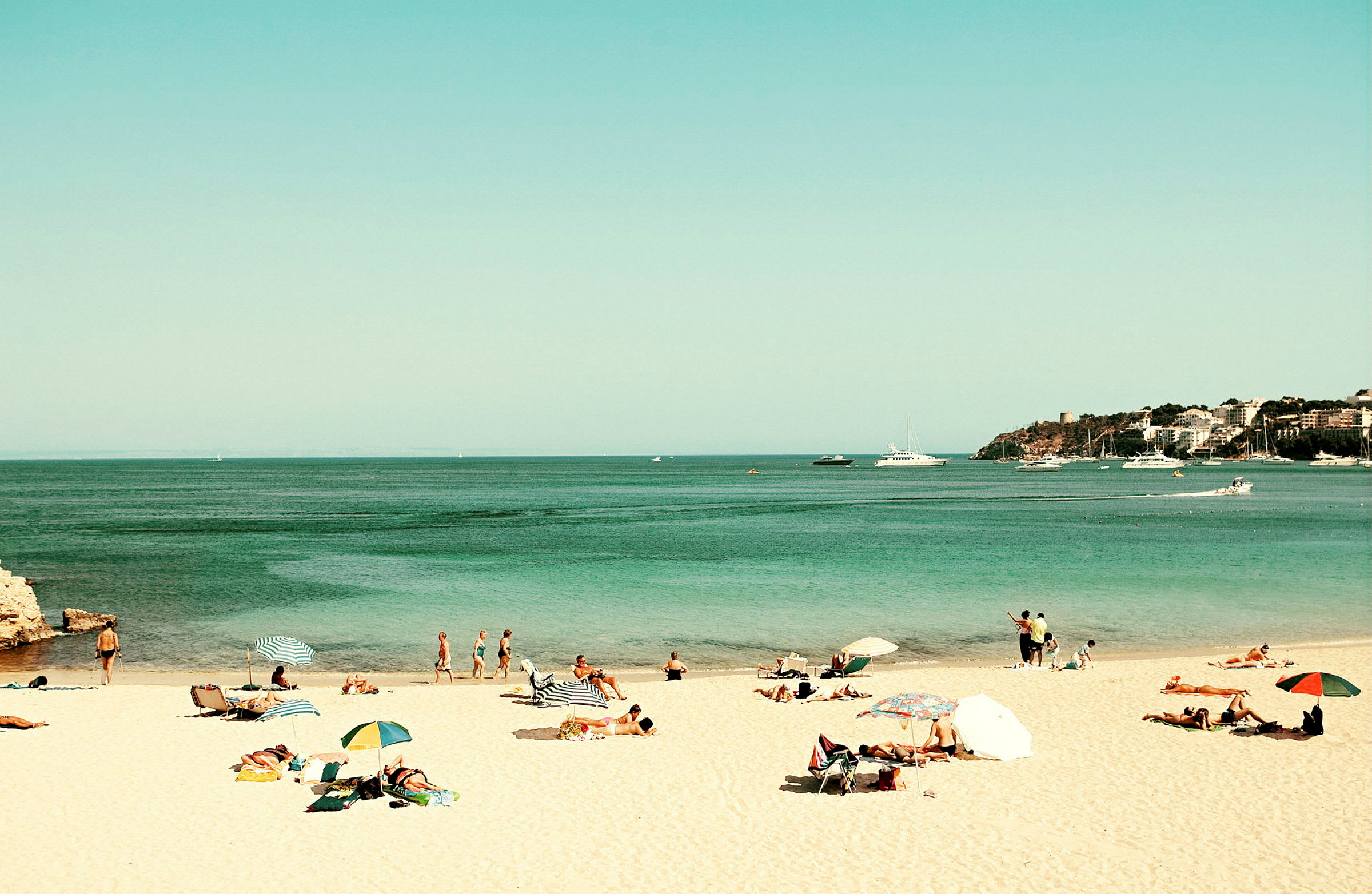 En strand med människor som solar och badar, parasoll och havet med båtar i bakgrunden. (AI-genererad text)