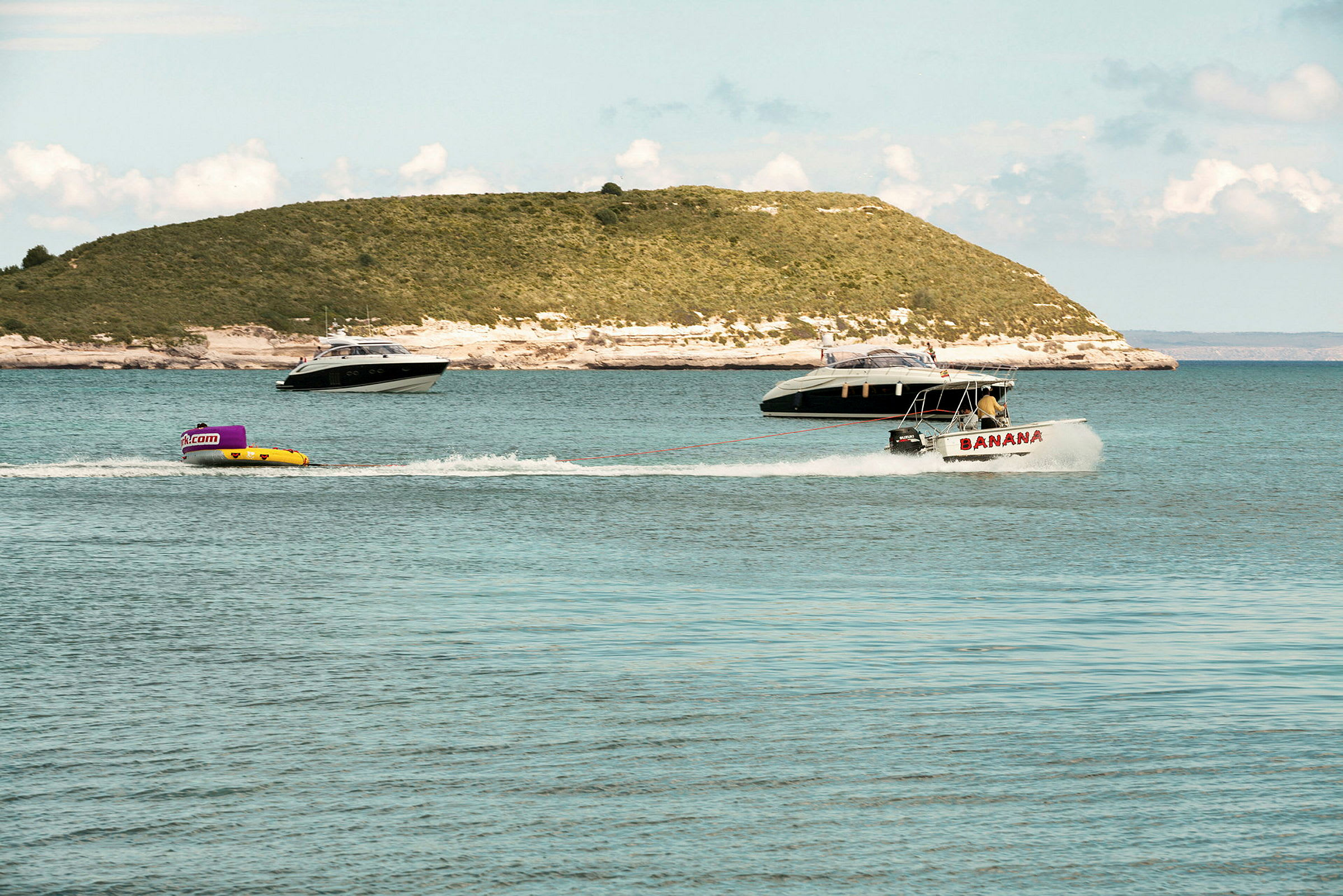 En motorbåt drar en uppblåsbar vattenleksak över havet. I bakgrunden syns två andra båtar och en grönskande ö. (AI-genererad text)