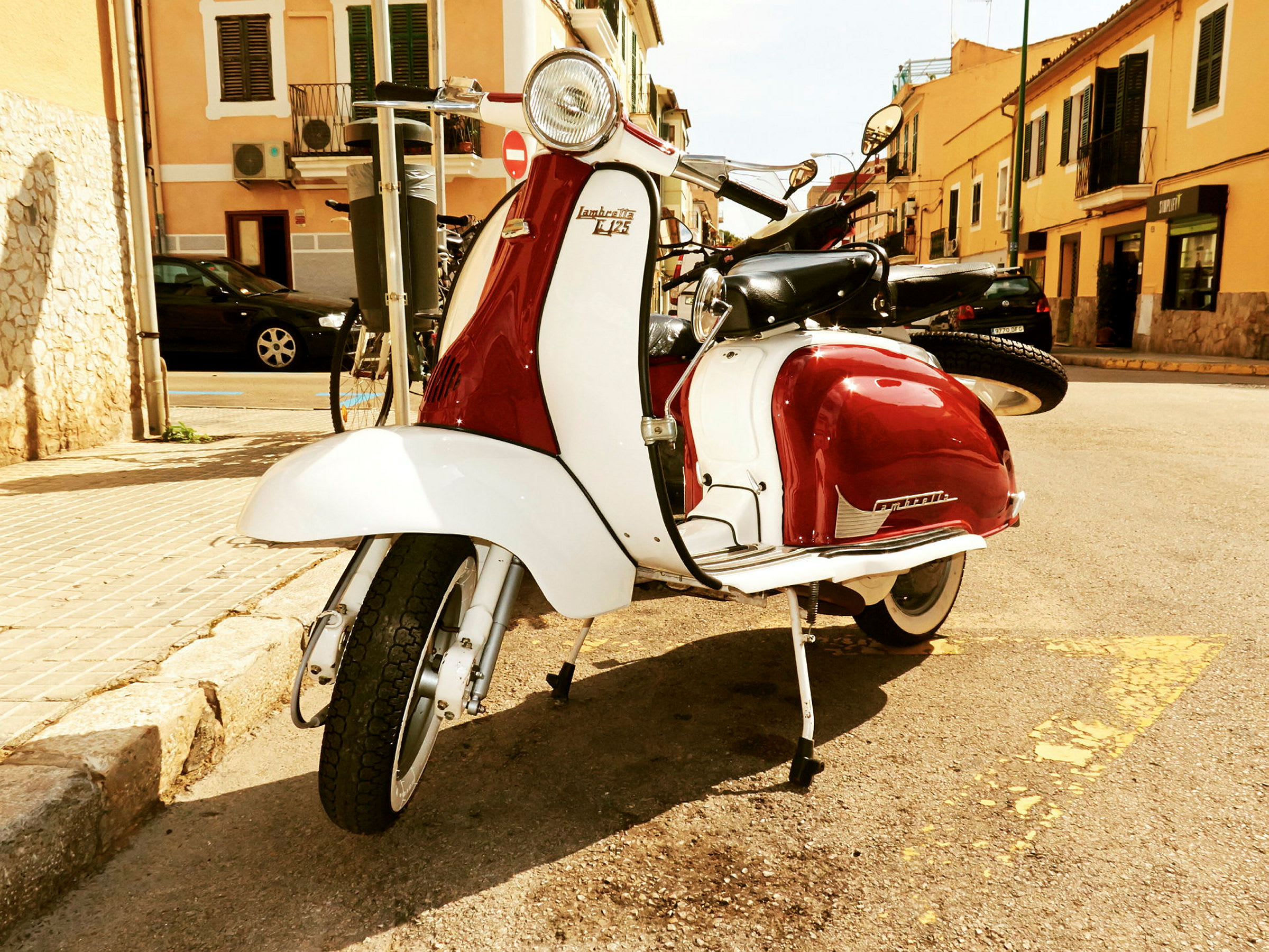 En klassisk röd och vit scooter är parkerad vid en trottoarkant på en solig dag. Bakgrunden visar byggnader och en gata. (AI-genererad text)