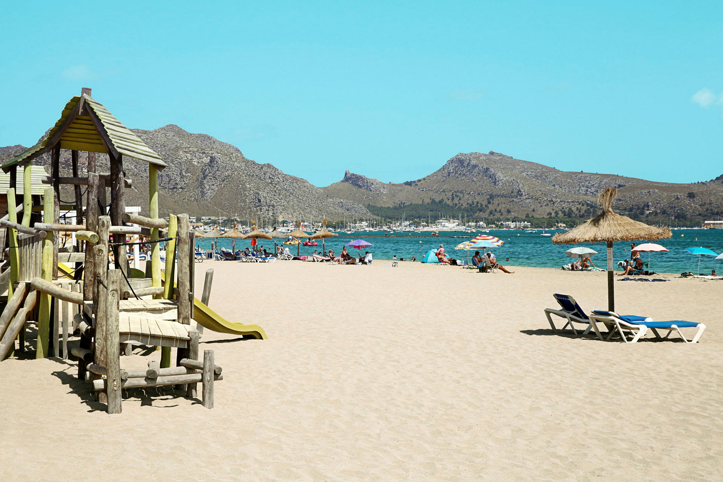 En sandstrand med lekplats, solstolar och parasoller vid vattnet med berg i bakgrunden. (AI-genererad text)