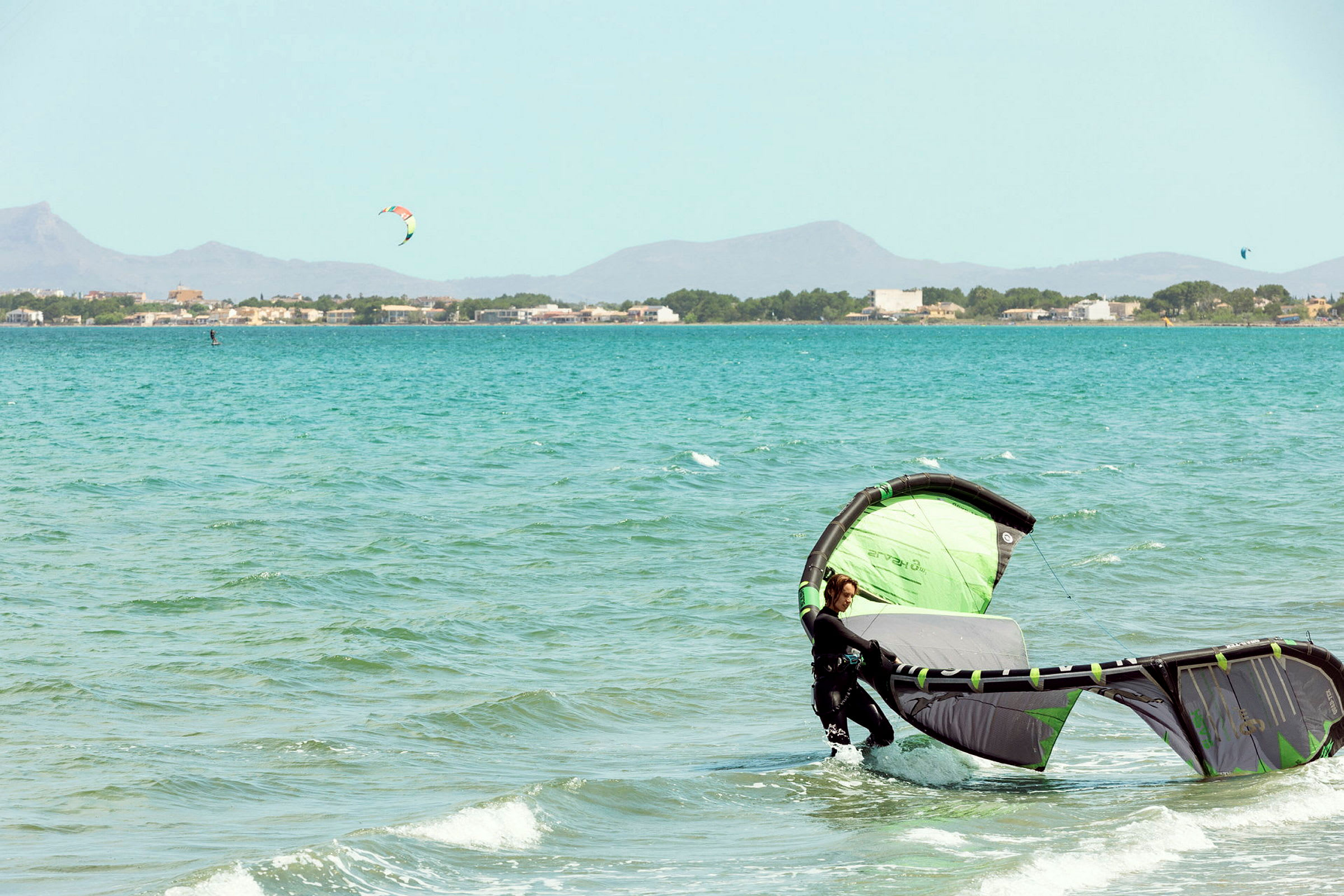 En person i vatten med en grön och svart kitesurfing drake, med en stad och berg i bakgrunden. (AI-genererad text)