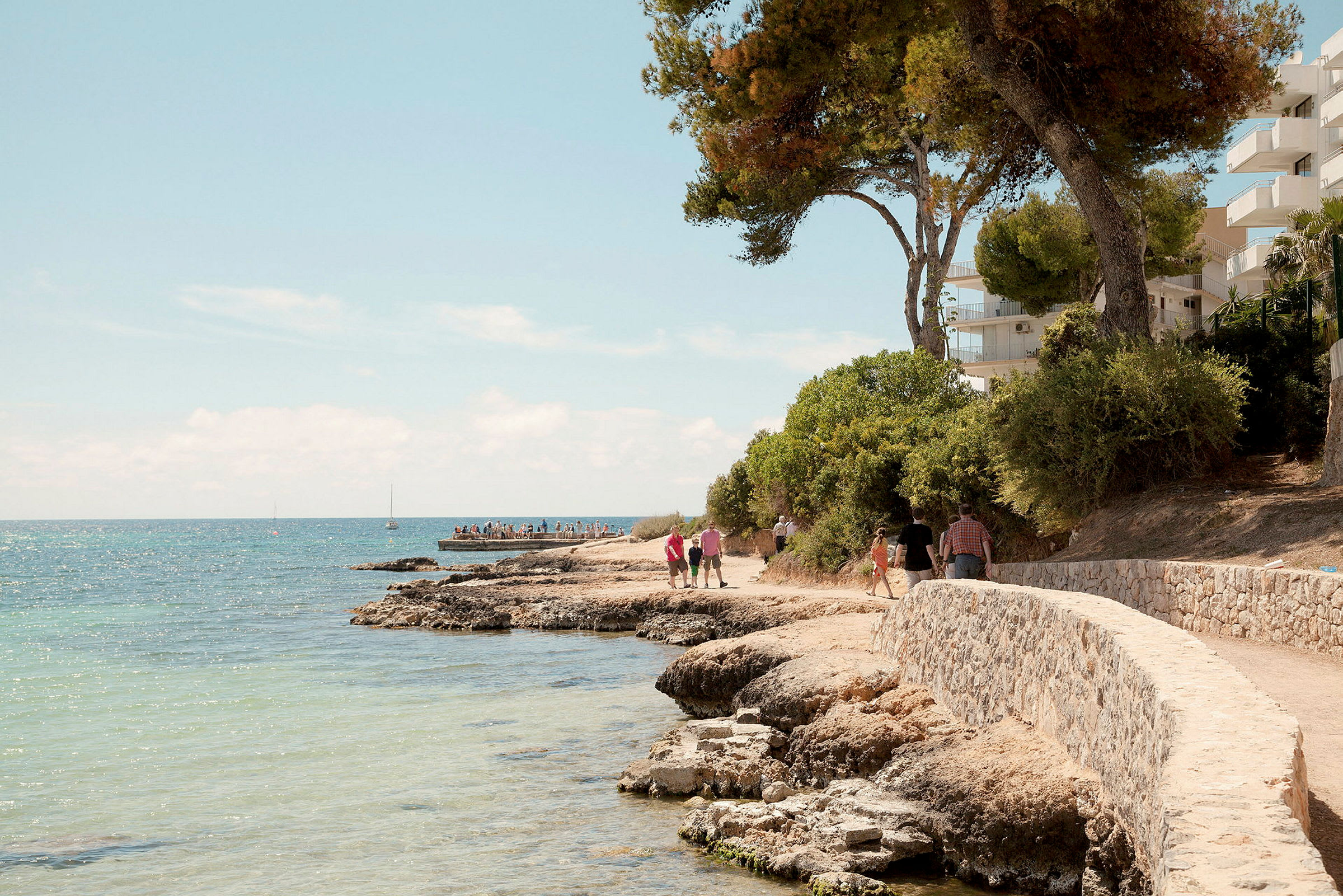 Stenstrand med slingrande gångväg intill havet, omgivet av träd och byggnader. (AI-genererad text)