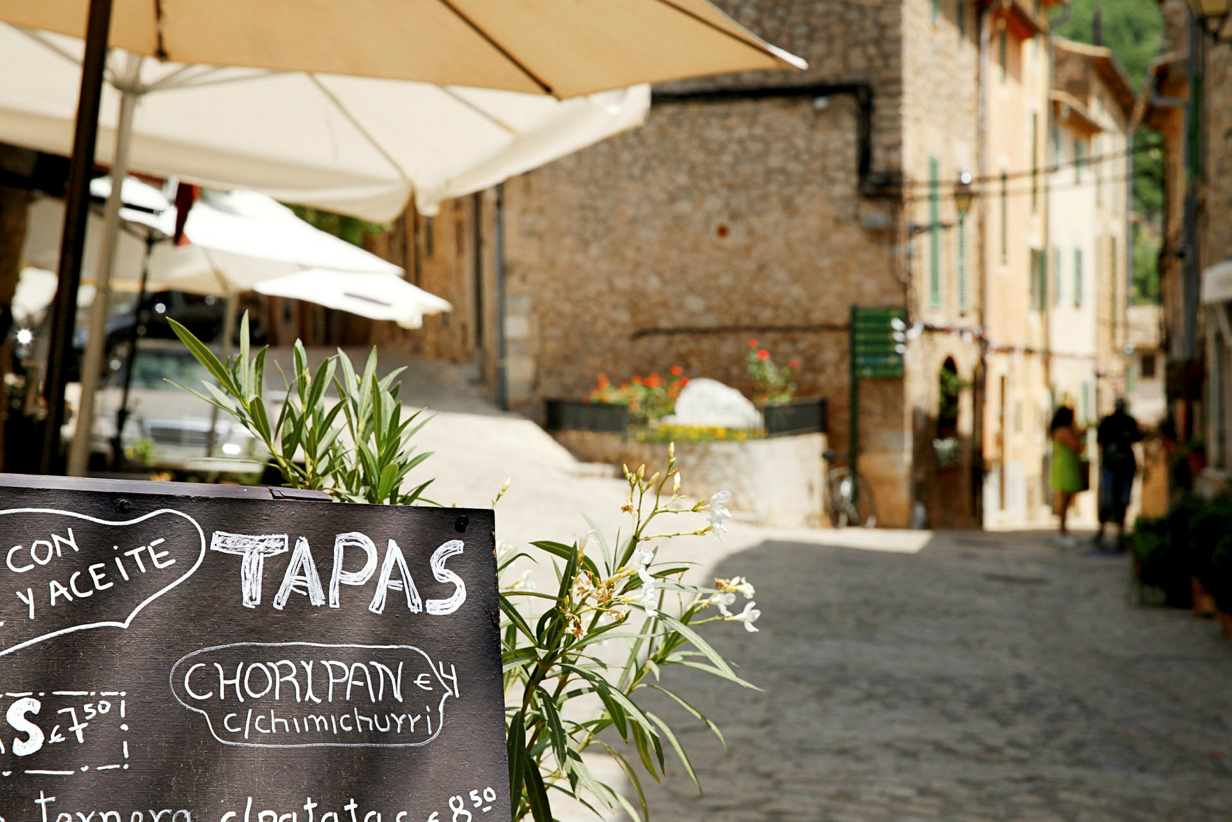 Gatuscen med ett svart menytavla med texten “tapas” i förgrunden och parasoll samt stenhus i bakgrunden. (AI-genererad text)
