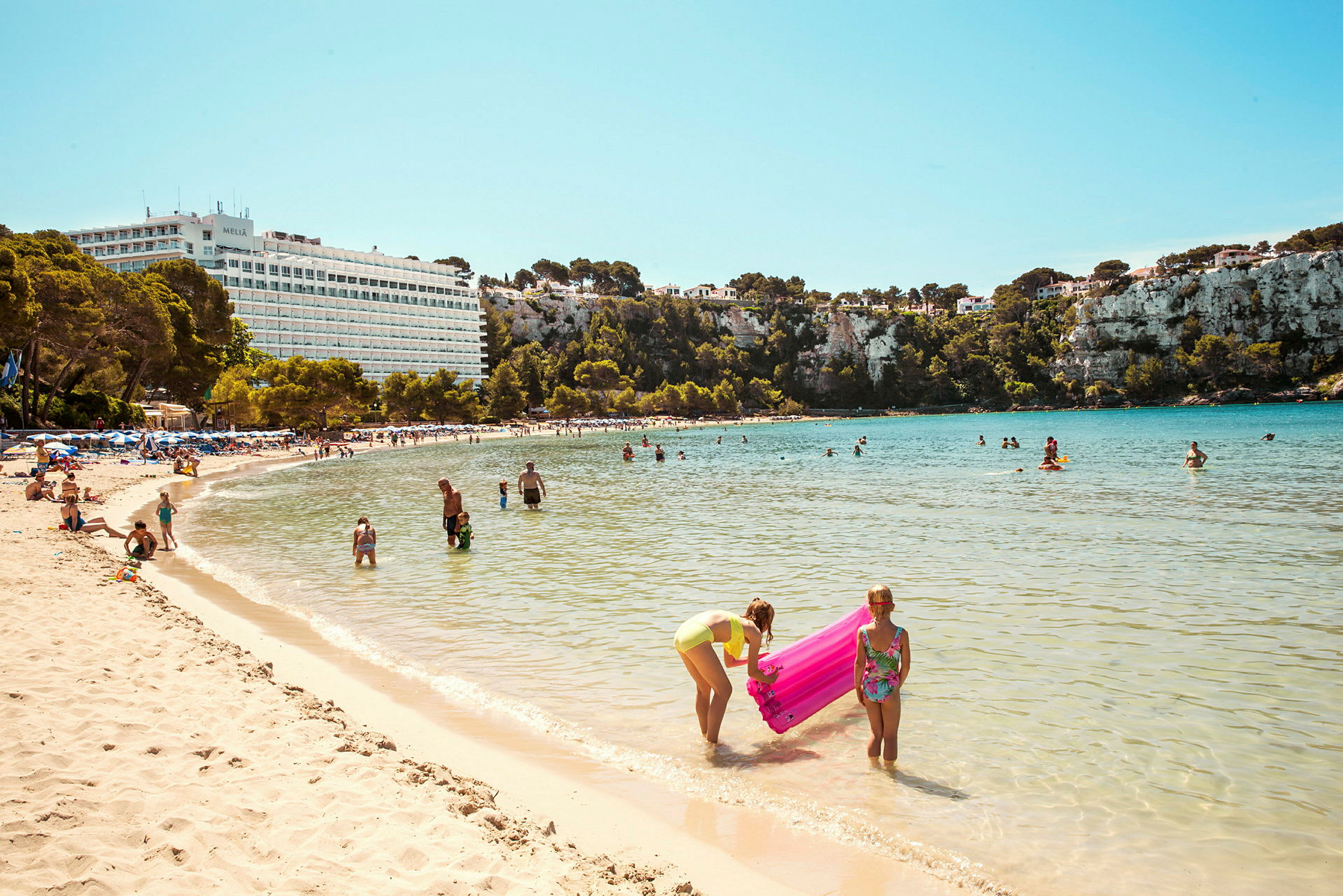 Strand med människor som badar och leker i vattnet framför en naturskön klippa och ett hotell. (AI-genererad text)