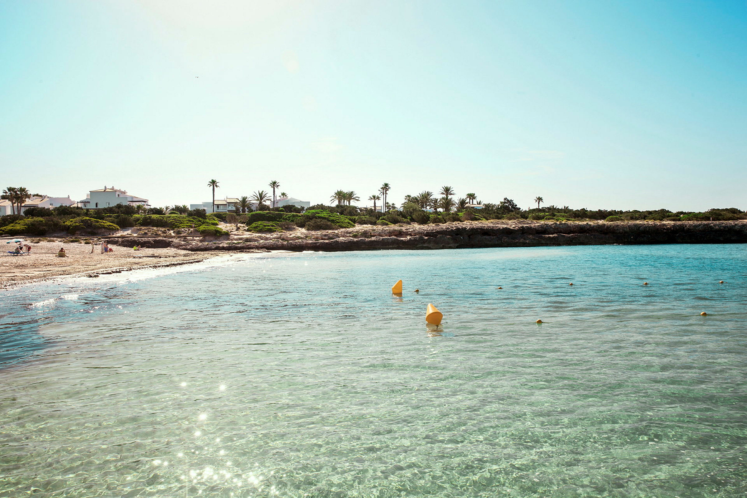 En strand med klart vatten, omgiven av gröna växter och hus i bakgrunden. (AI-genererad text)
