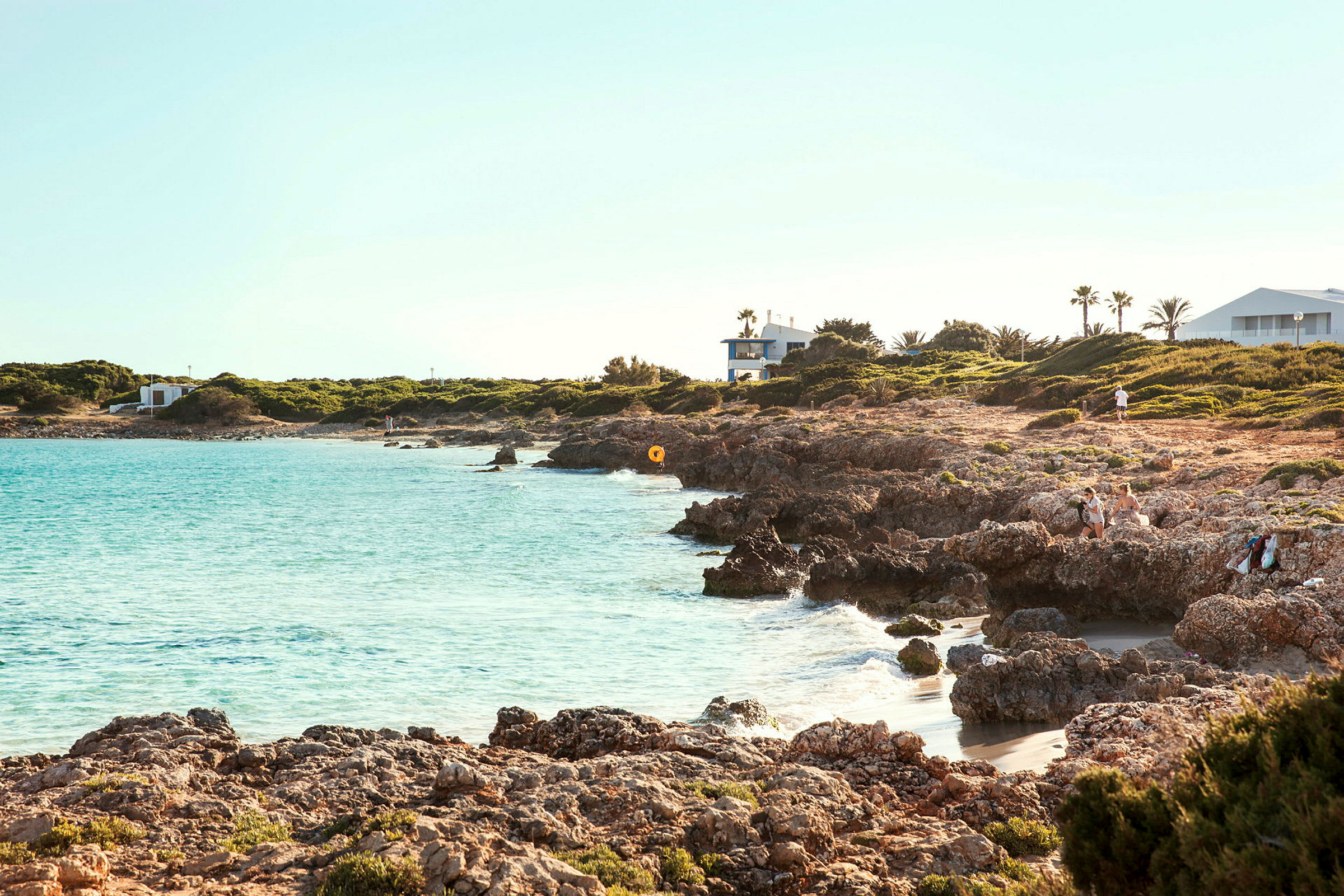 Strand med klart vatten och klippigt landskap, omgivande grönska och byggnader i bakgrunden. (AI-genererad text)