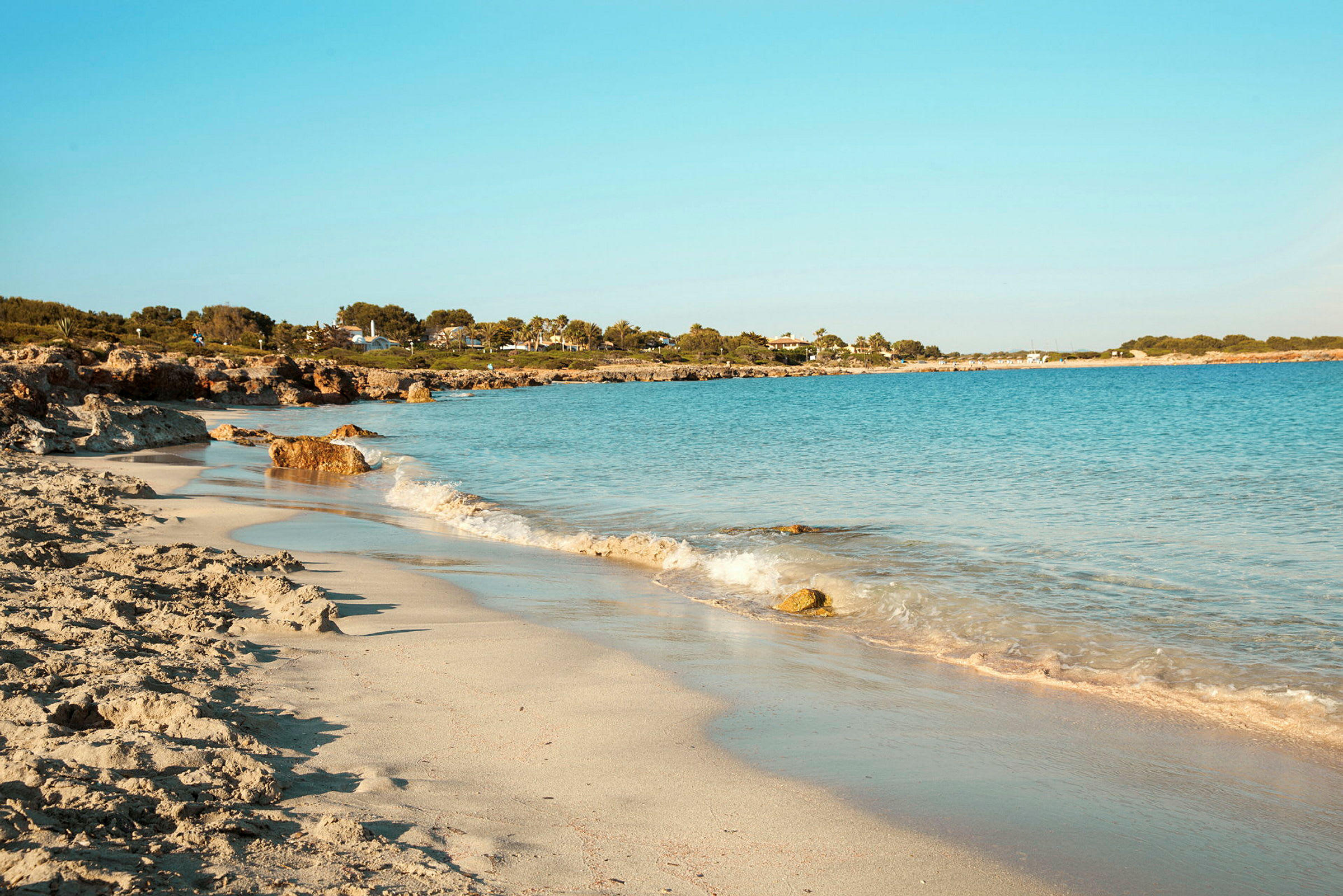 En strand med ljus sand och blått hav, omgiven av grönska och byggnader i bakgrunden. (AI-genererad text)