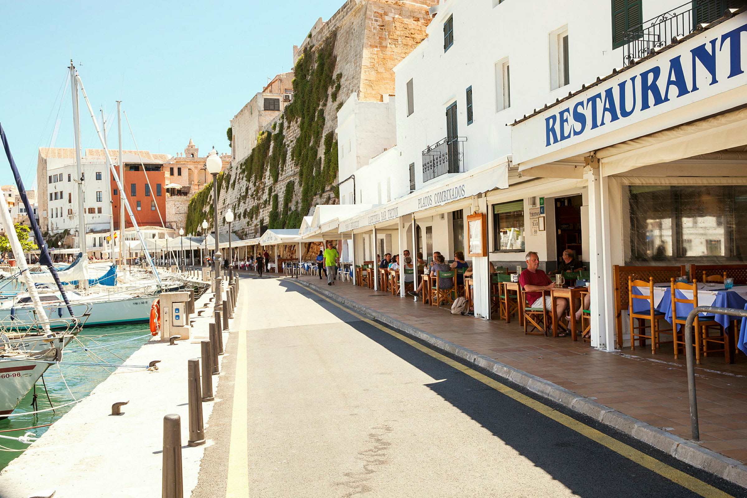 En hamnpromenad med restauranger och båtar vid kajen. Det är en solig dag med människor som sitter utomhus på restaurangernas terrasser. (AI-genererad text)