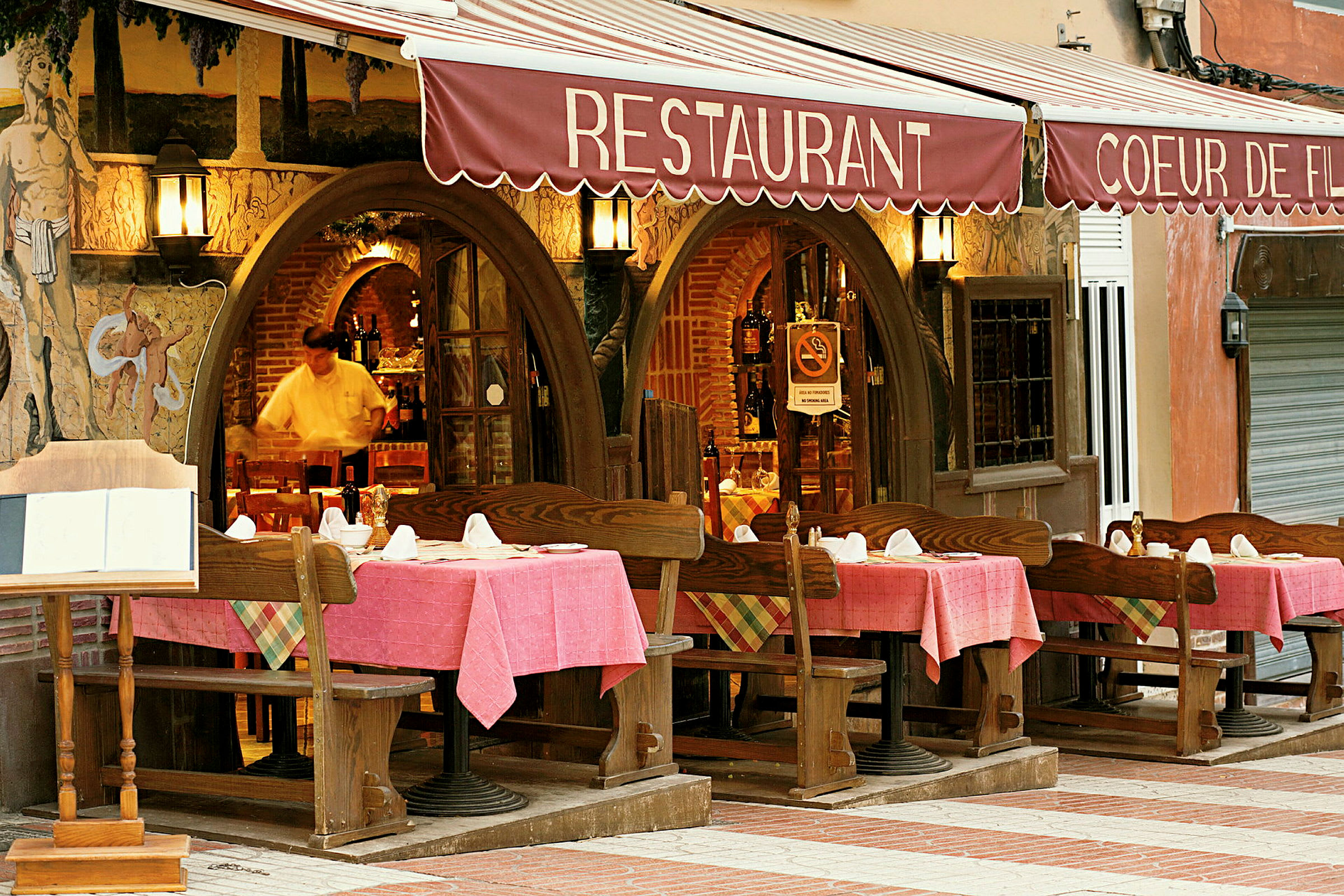 Entré till restaurang med bord och stolar utomhus, dukade med rosa dukar. (AI-genererad text)