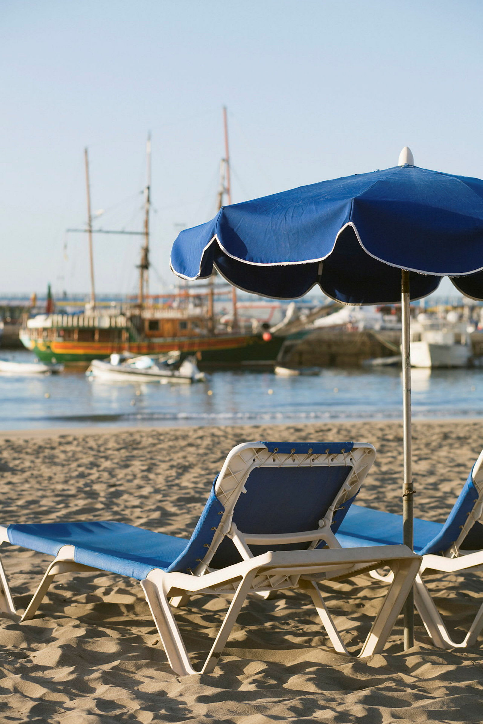 Två solstolar med blå dynor och ett blått parasoll på en sandstrand med segelbåtar i bakgrunden. (AI-genererad text)