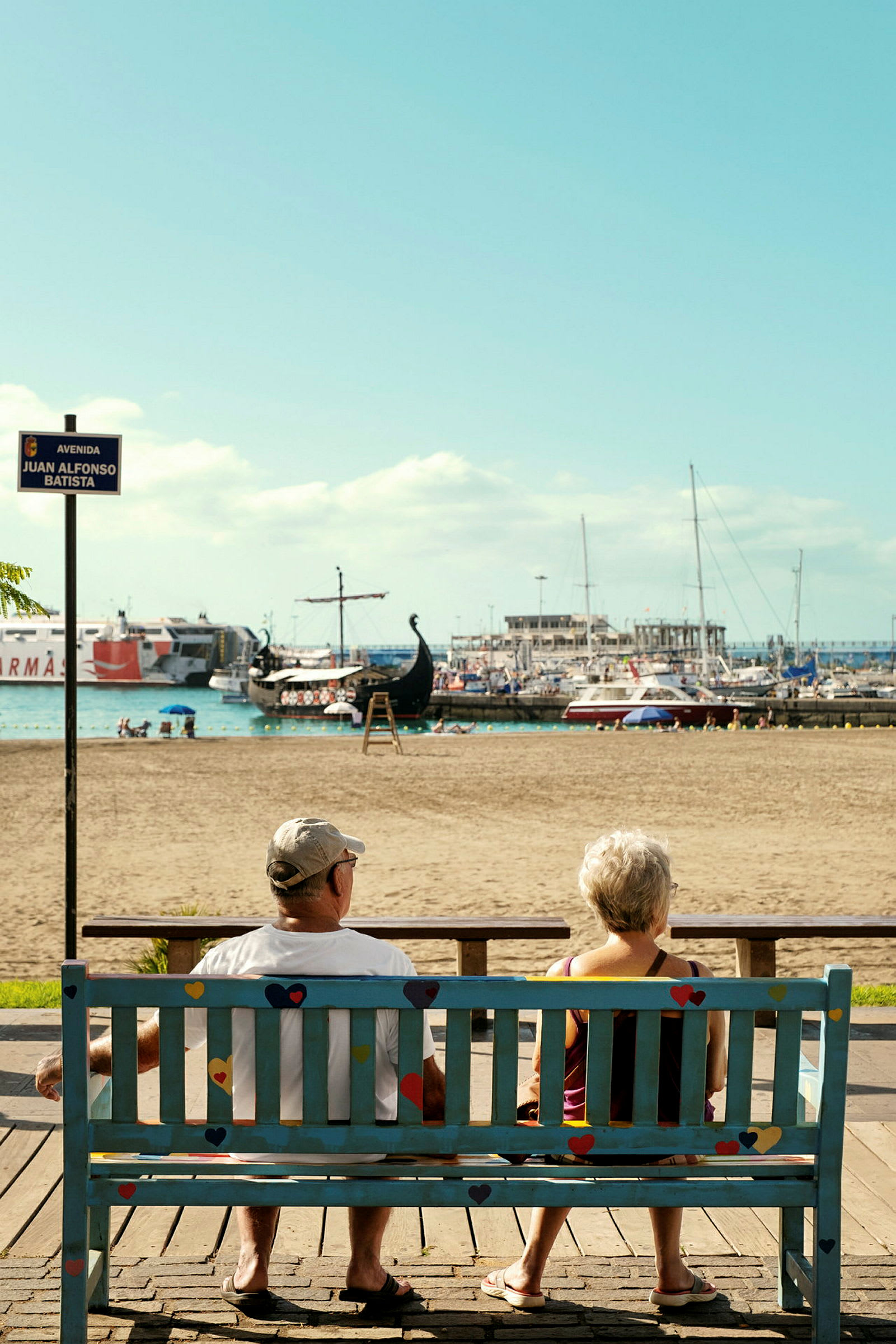 Två personer sitter på en bänk med utsikt över stranden och hamnen. (AI-genererad text)